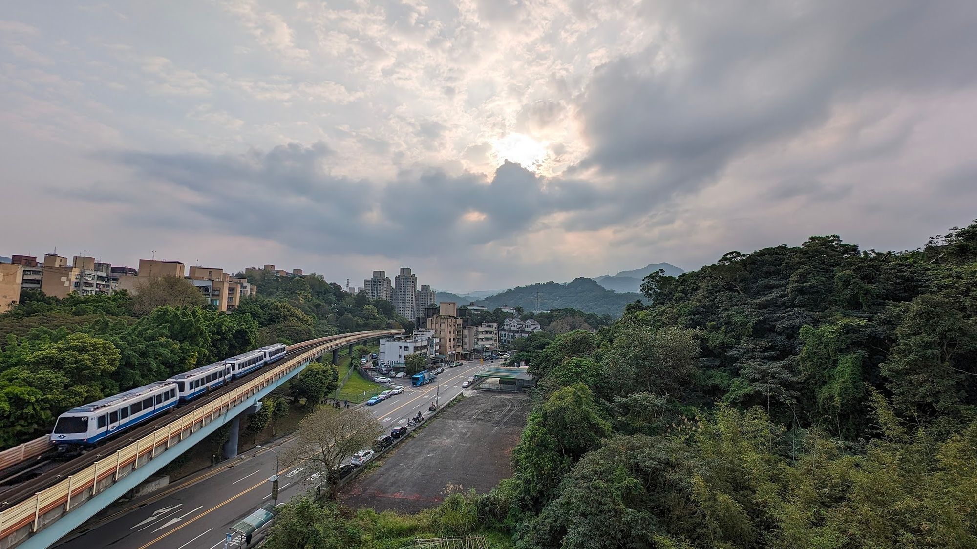 Blick aus meinem Schlafzimmerfenster. Rechts Dschungel, zentral eine Strasse und Hochbahnstrecke, links Baeume und dahinter Wohnhaeuser. Im Hintergrund sieht man dicht bewaldete Berge