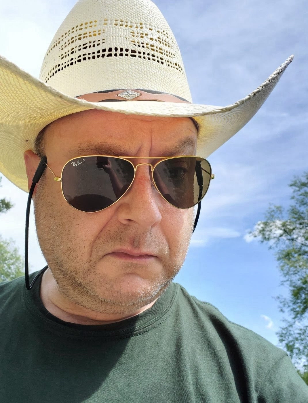 A devilishly handsome man with a pale cowboy hat, Rayban sunglasses, a green T-shirt, looking grumpy against a blue sky.