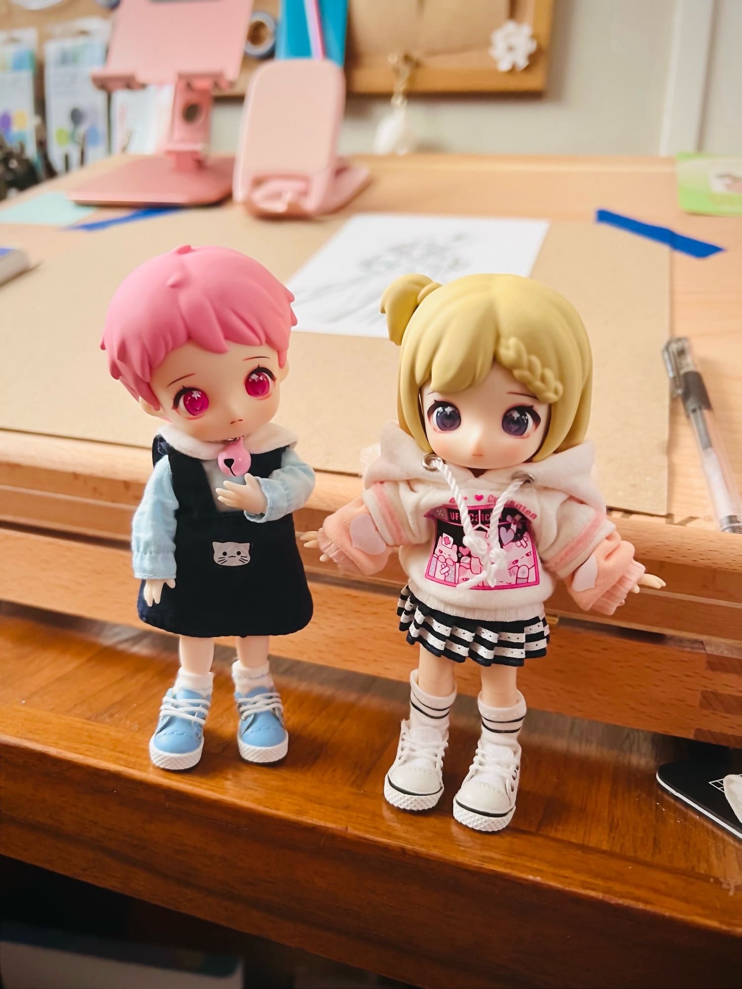 Two small Obitsu11 dolls. One has short pink hair and a dark blue dress with blue shirt and shoes. The other has a blonde hair and a side pony in a tiny hoody and pleated skirt.