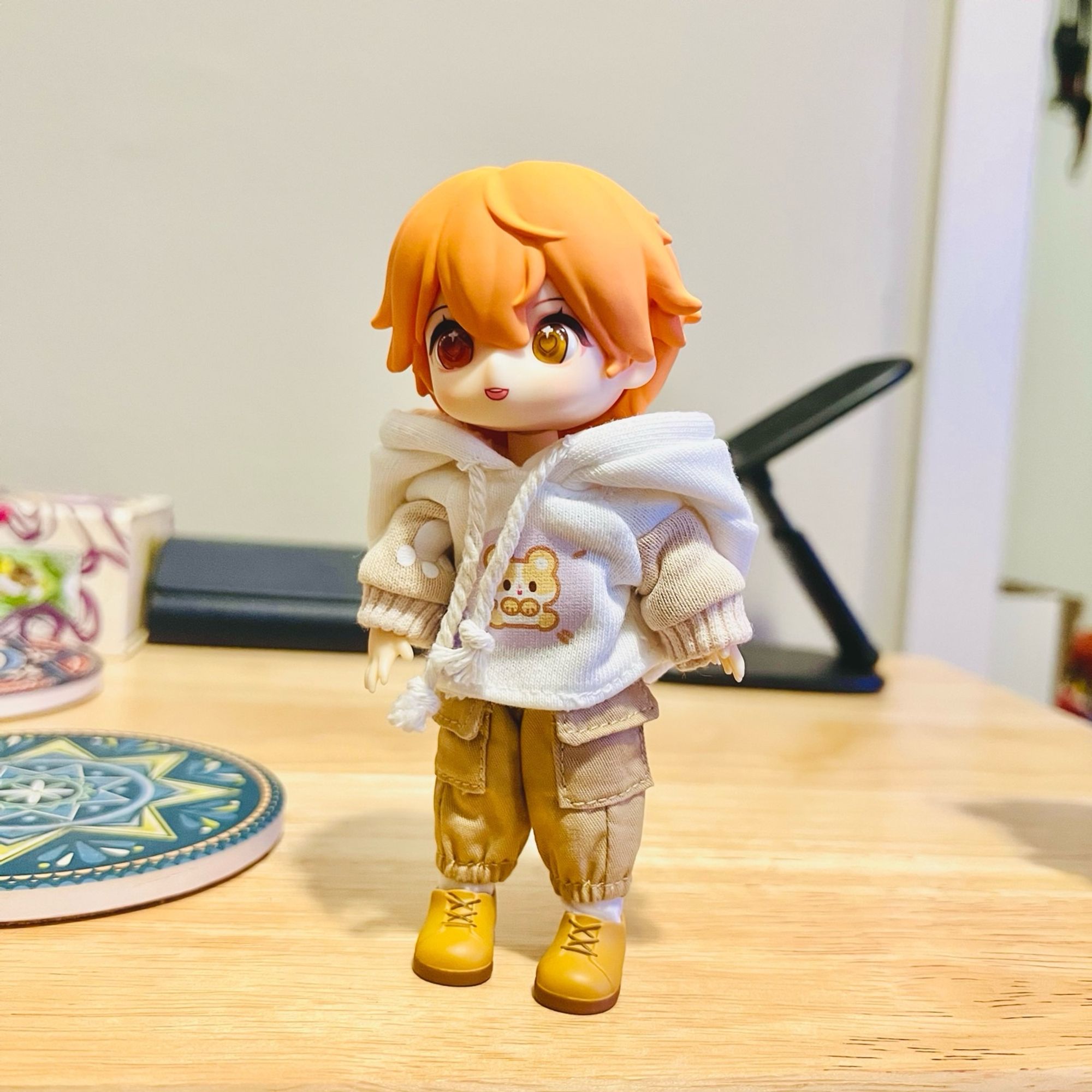 A photo of an Obitsu11 ball jointed doll with orange-red hair wearing a hamster-print hoodie, dark khaki cargo shorts, and little yellow shoes. He stands on a wooden surface in front of a coaster, a case for eyeglasses, and a phone stand.