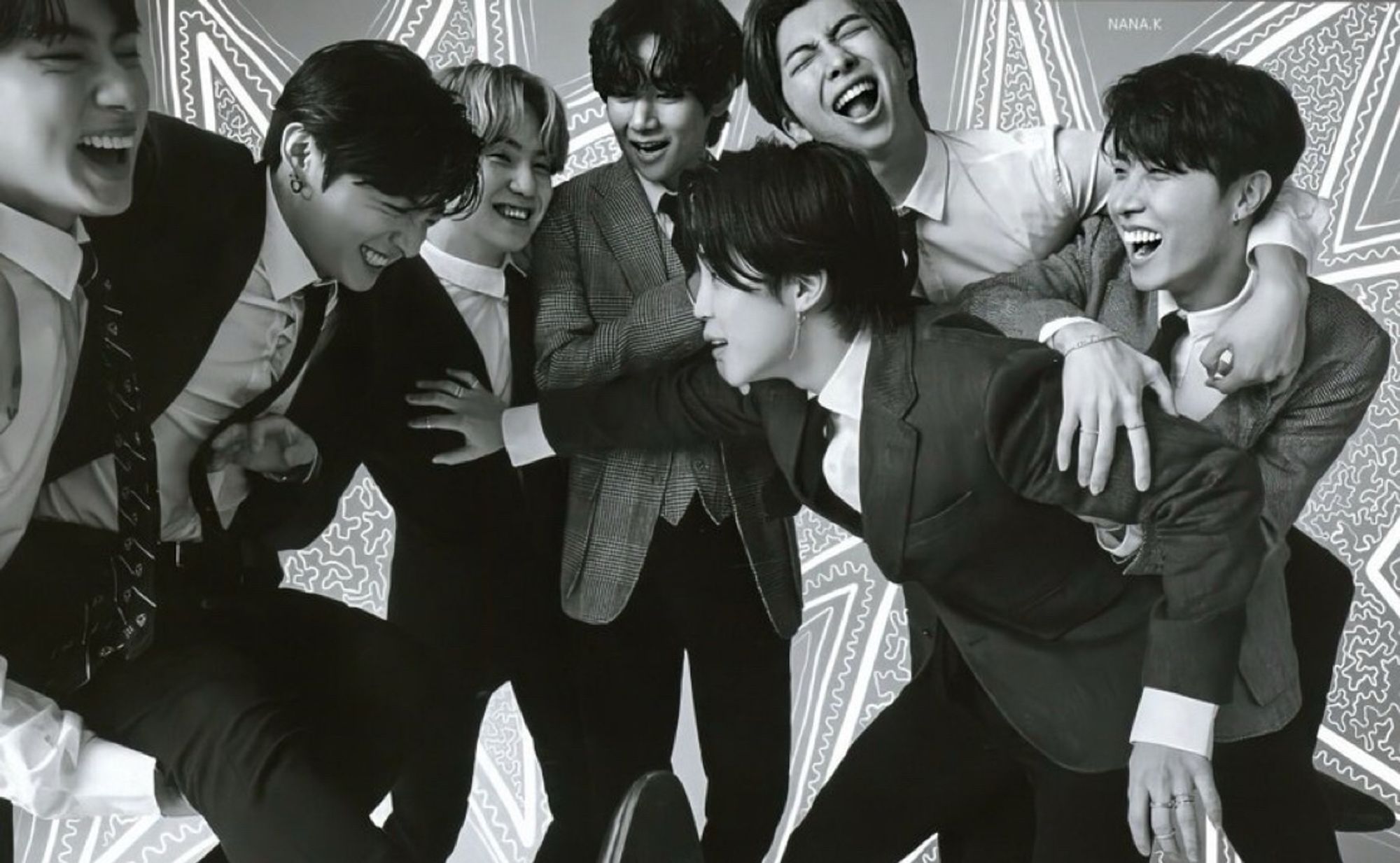 Bts in suits crowded around each other laughing; jimin and jk appear to be messing around. Photo in black and white.