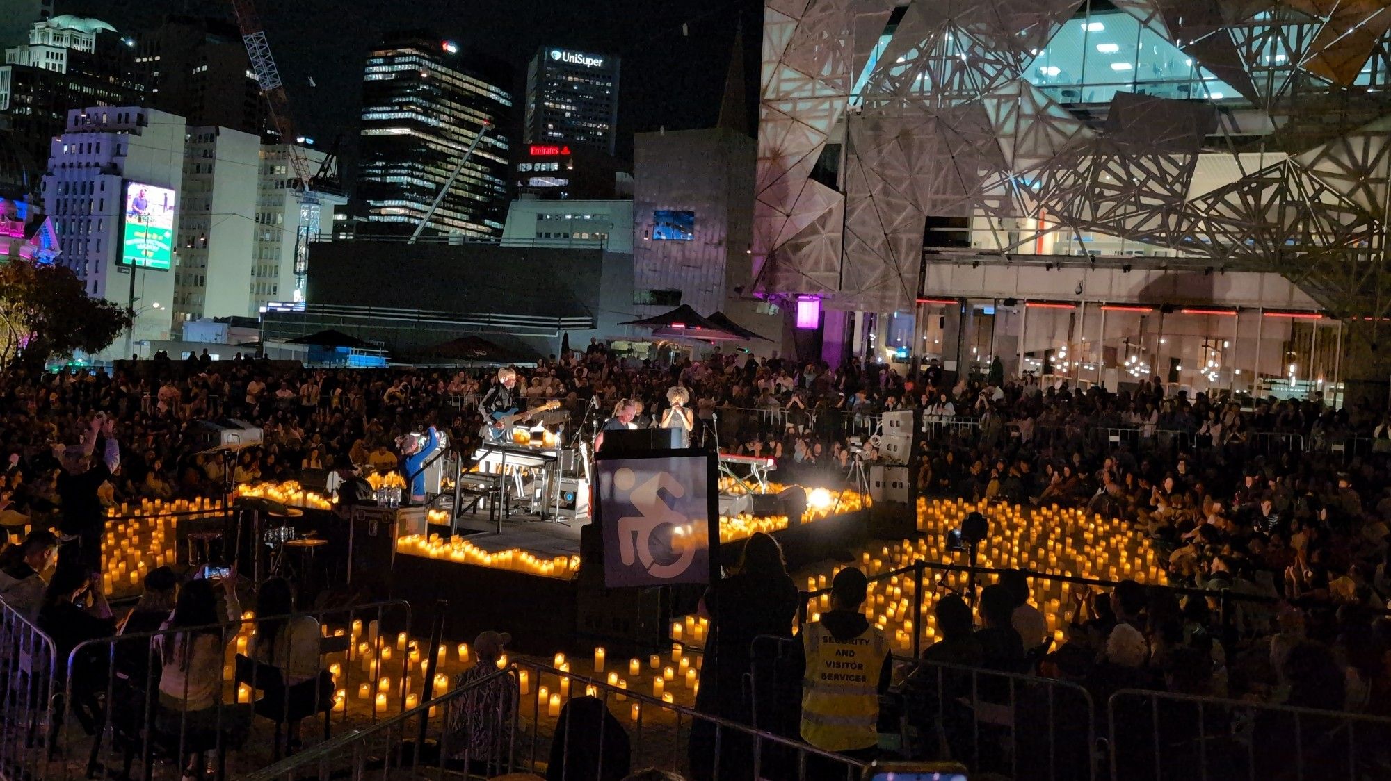 Candlelight concert at Federation Square. There is a band performing on stage surrounded by candles and an audience