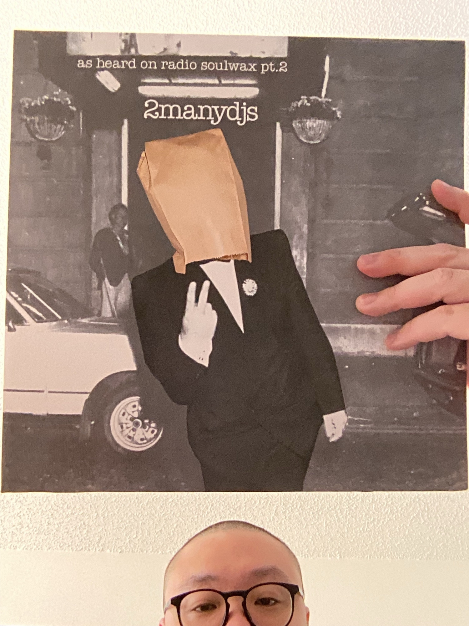 The cover for 2manydjs’ As Heard On Radio Soulwax Vol. 2, a man in a tux with a paper bag over his head holding two fingers up in front of the entrance to a club