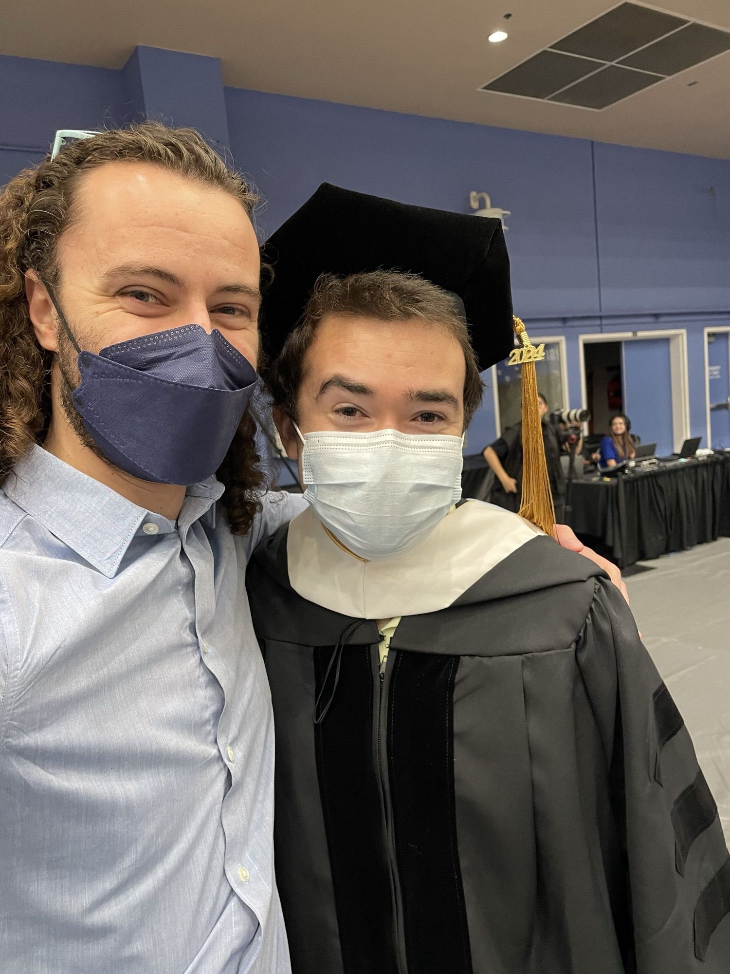 Brandy Juniper Lewis graduating in her PhD robes, standing with her partner and both wearing masks.