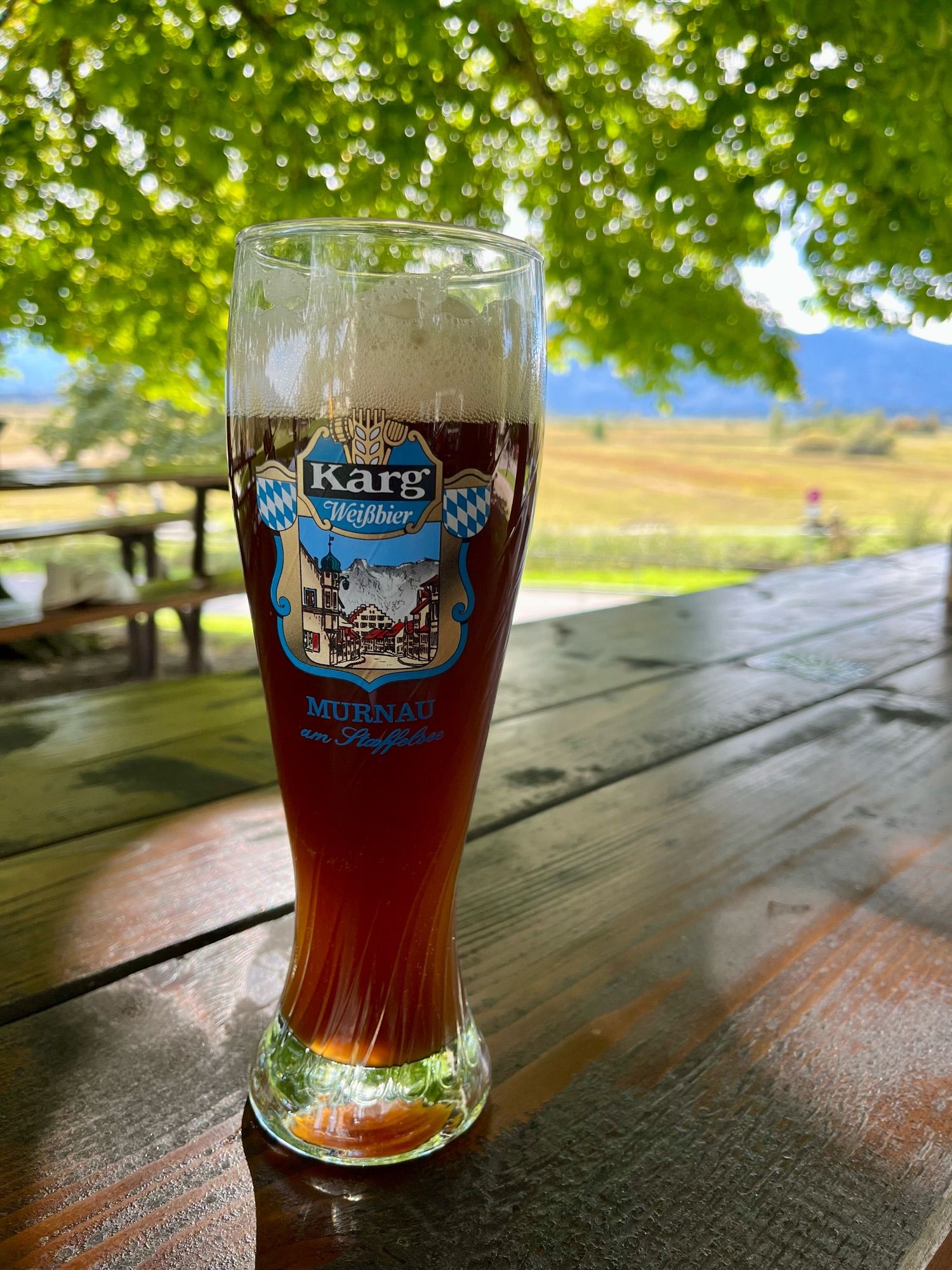 Auf einem Holztisch steht ein dunkles Weißbier aus der Murnauer Brauerei Karg. Im Hintergrund das Murnauer Moos.