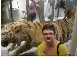 Julie, a white person with short brown hair wearing glasses and a bright green shirt, poses next to a taxidermied tiger.