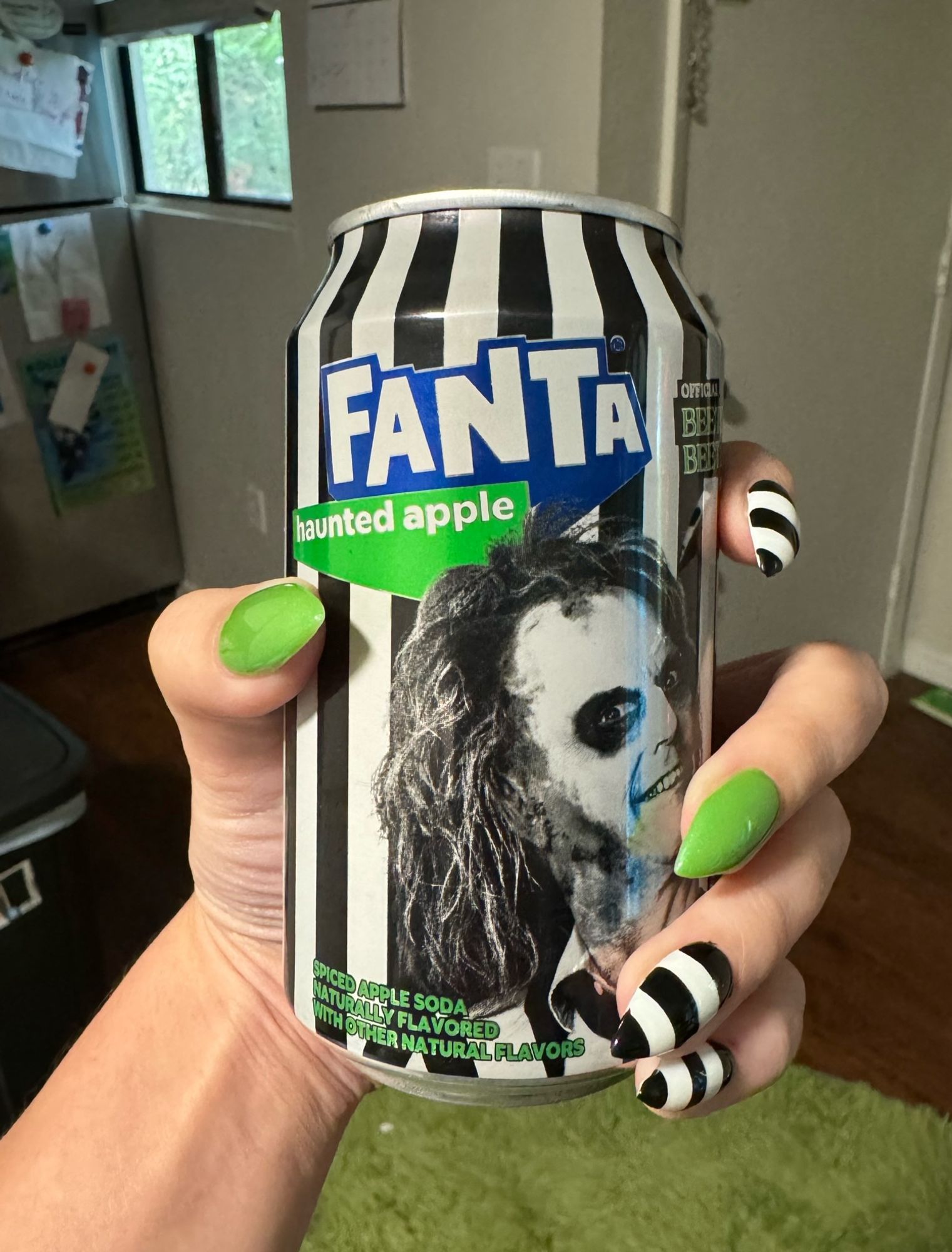 my hand holding up a black and white striped fanta can with beetlejuice on it and  “haunted apple” flavor in green, matching my nails exactly lmao, they’re pointy glittery green thumb and middle finger, black and white striped on the others