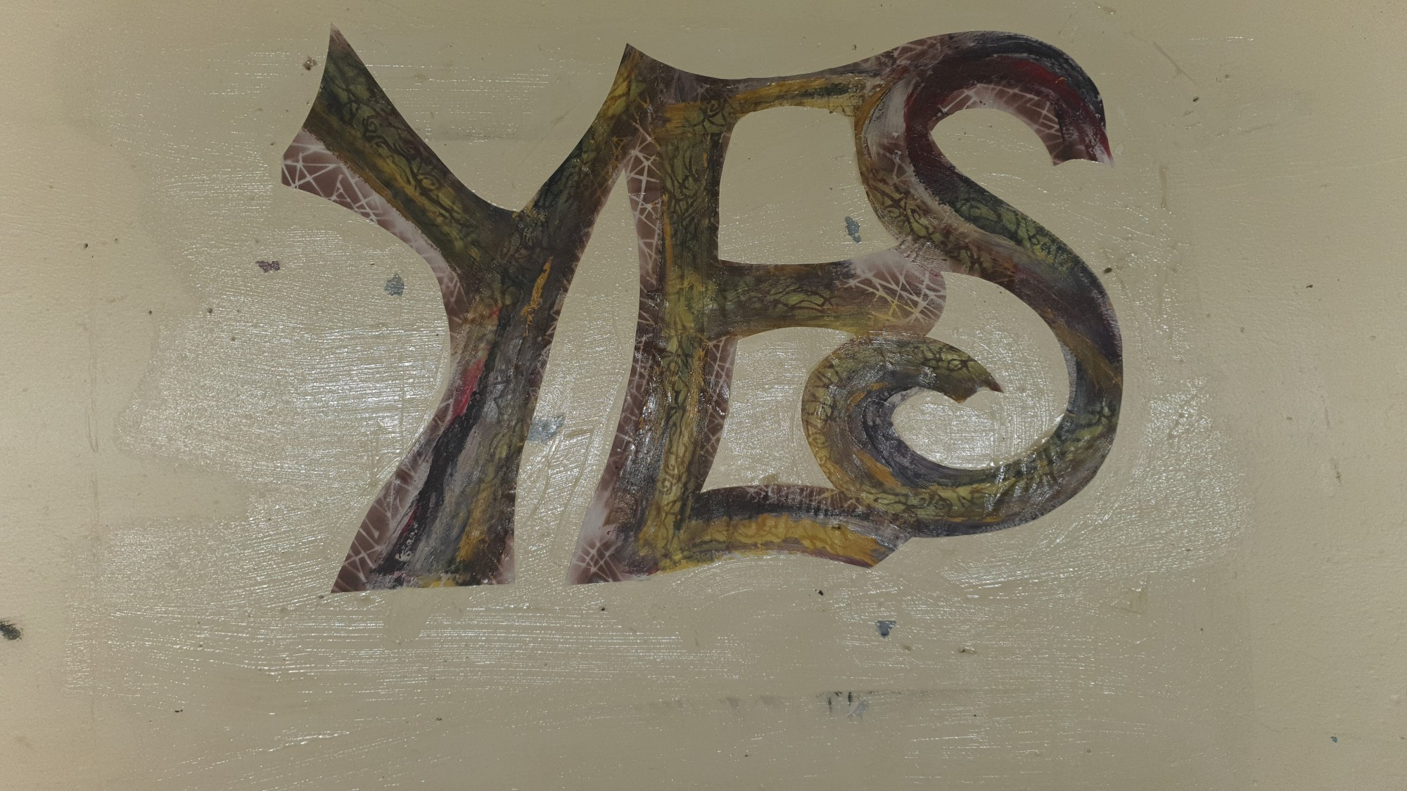 The word 'Yes' is pasted up on the wall inside a Canberra bus shelter. The letters have been cut out of paper that has been painted to resemble wood. The colours are different shades of brown to yellow, with little bits of white cross hatching that look like peeling bark.