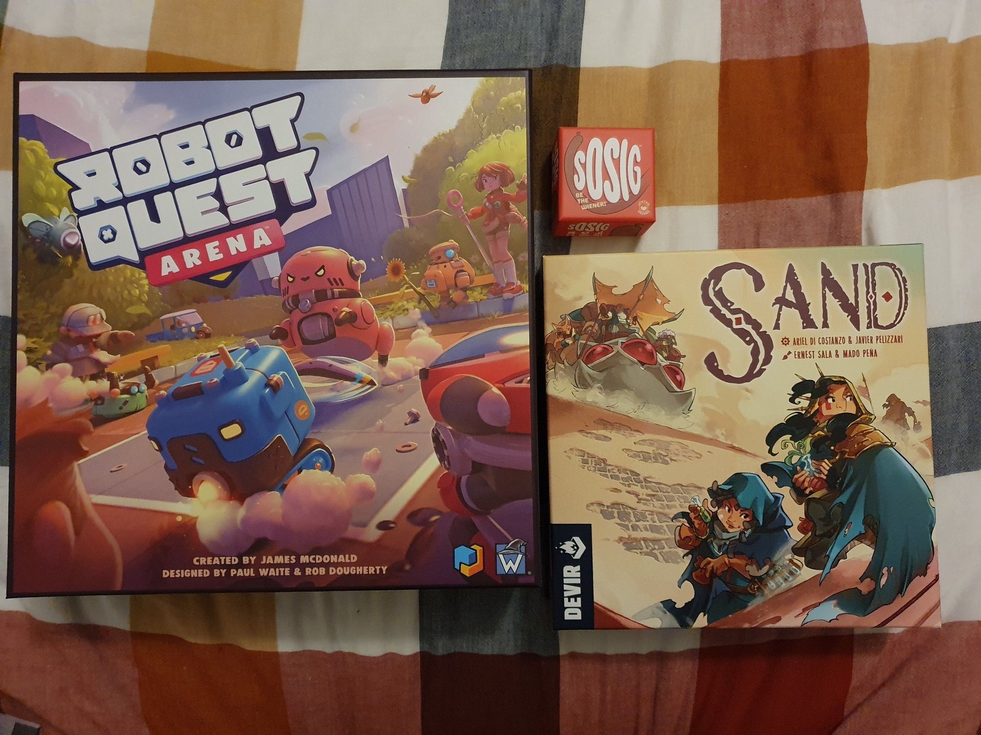 a photo of three boxed boardgames, from left to right: robot quest arena, sosig and sand