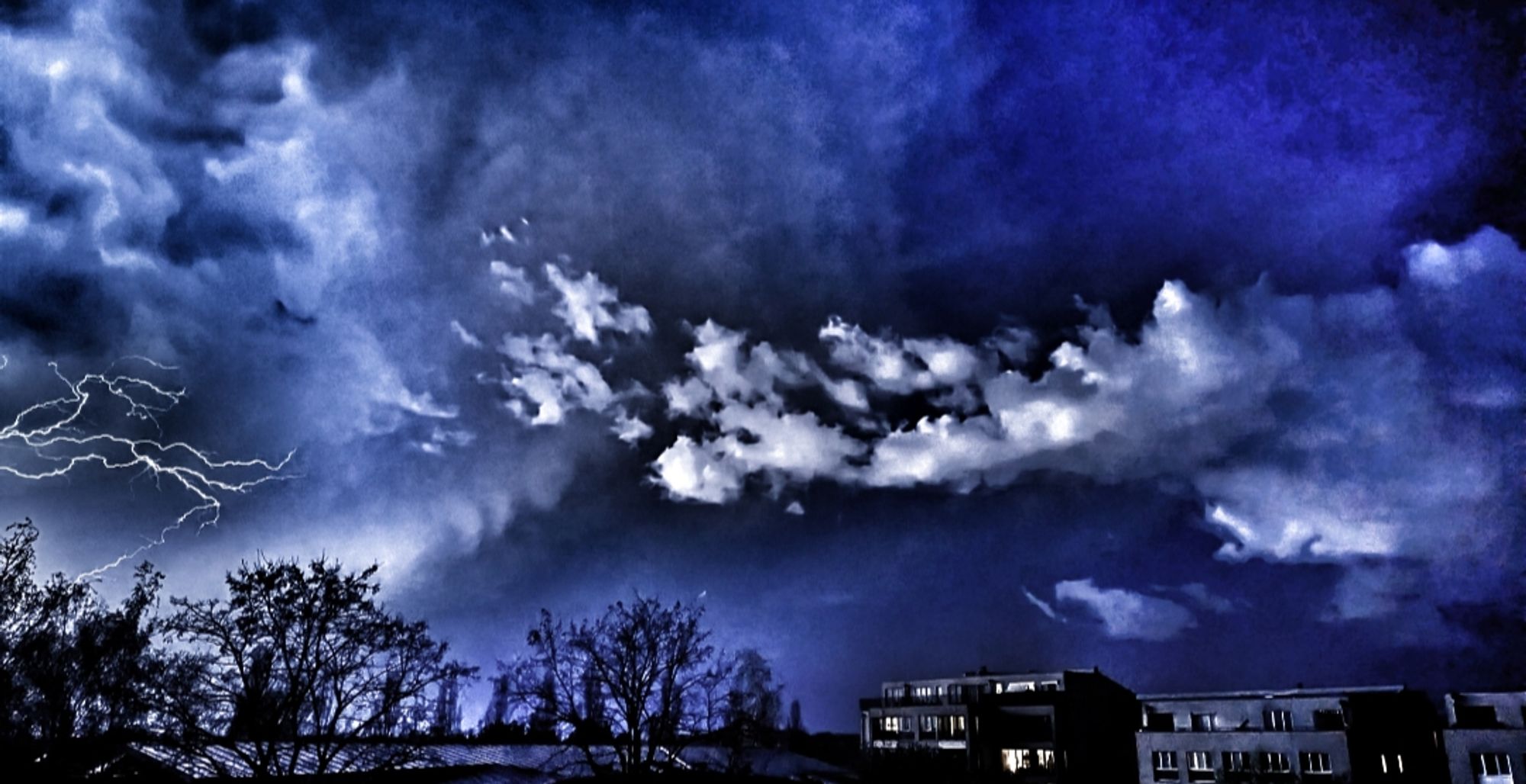 Aufziehendes, schweres Unwetter über einer Stadt. Blitzer und Wolken, zerrissener Himmel.
Einige erleuchtete Fenster...