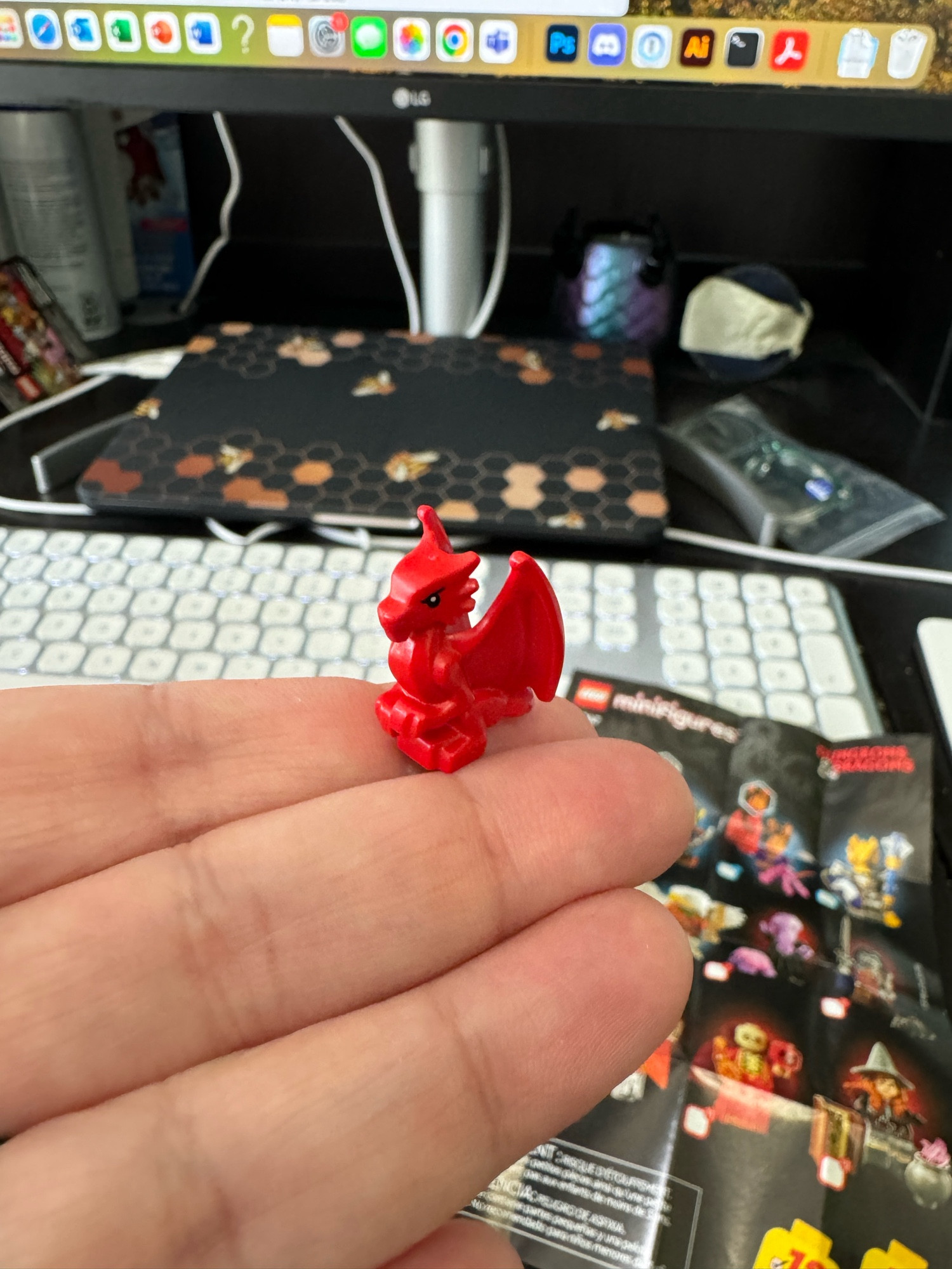 My fingers, with hand palm up in front of a messy desk, with a tiny red Lego dragon perched on the tip of my forefinger. It’s very small and very cute!