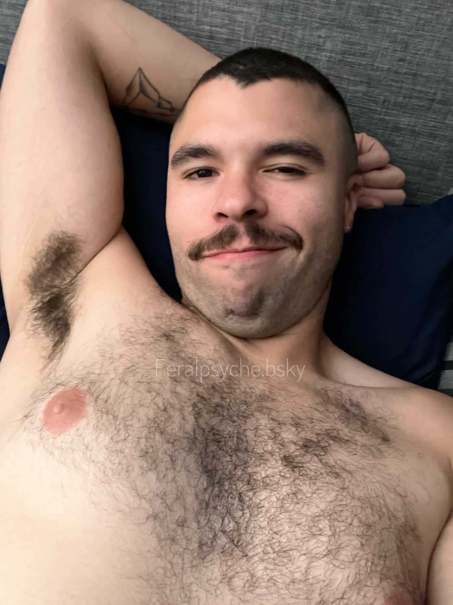 A hairy, shirtless guy, visible from his nipples up, laying in bed with an arm behind his head, exposing a hairy armpit