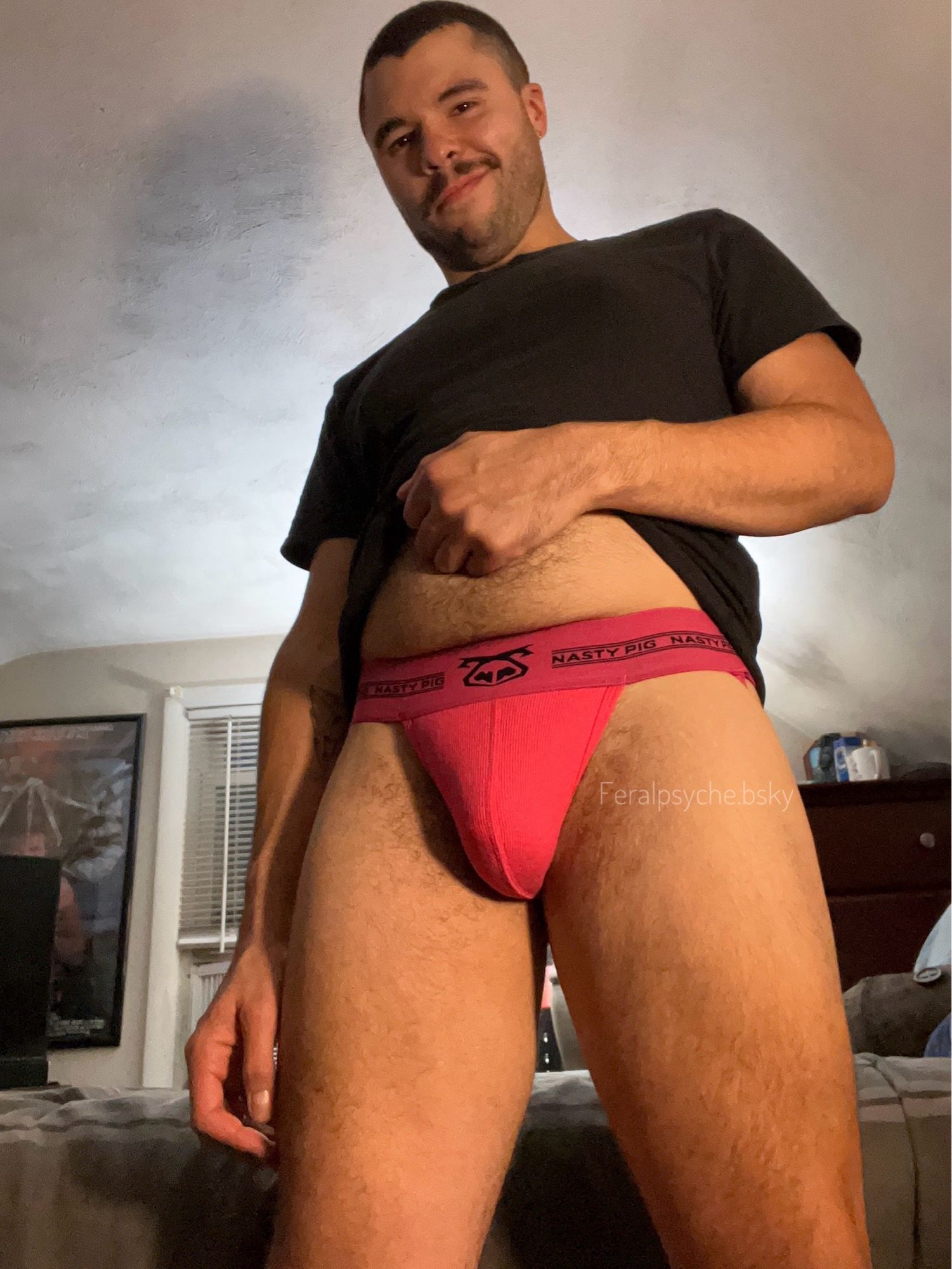 A guy in a black shirt and a pink jock, slightly lifting his shirt to show his hairy tummy