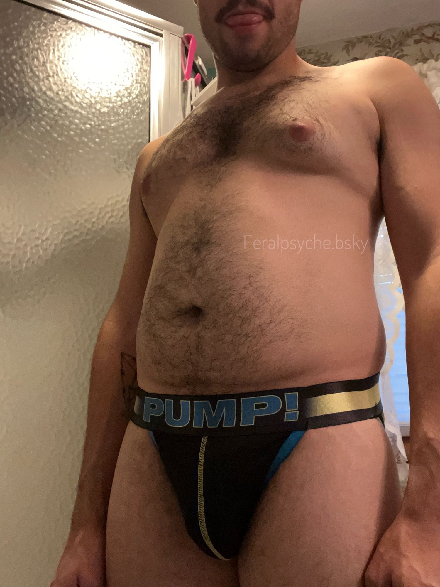 A shirtless, hairy guy wearing a blue and yellow Pump! jockstrap, standing in his bathroom.