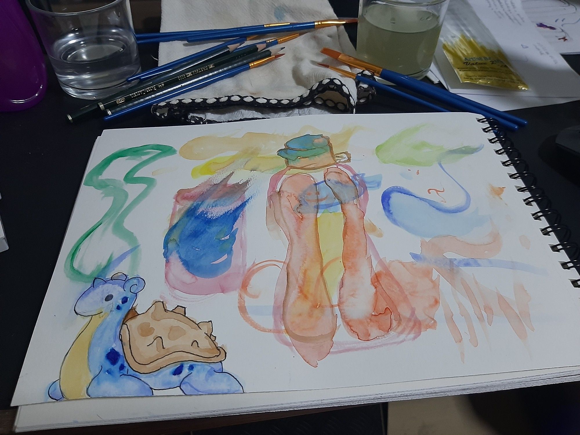 Mesa com vários pincéis, dois copos de água, lápis e um caderno. Há vários teste de tinta de aquarela e pincel na página mostrada, um estudo de uma garrafa um desenho pintado do Pokémon Lapras. É uma página de iniciantes em aquarela