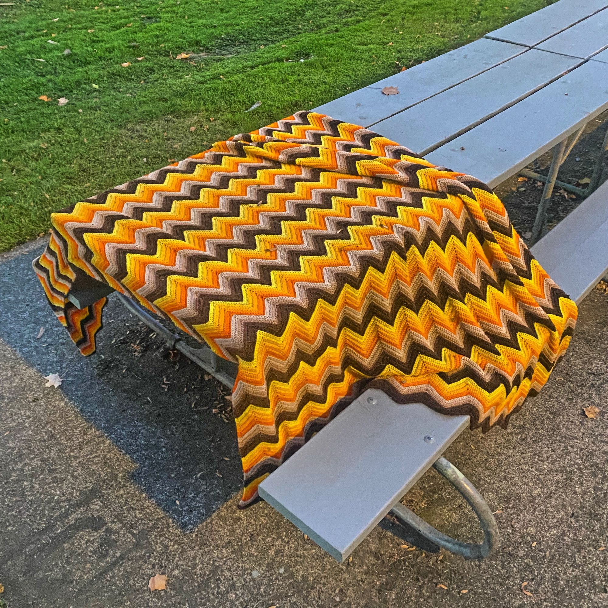 A crocheted afghan spread out across a picnic table. It is made up of stripes of yarn in a ripple pattern in “earth tones.” Such an afghan was a very popular design in the USA in the ‘seventies.