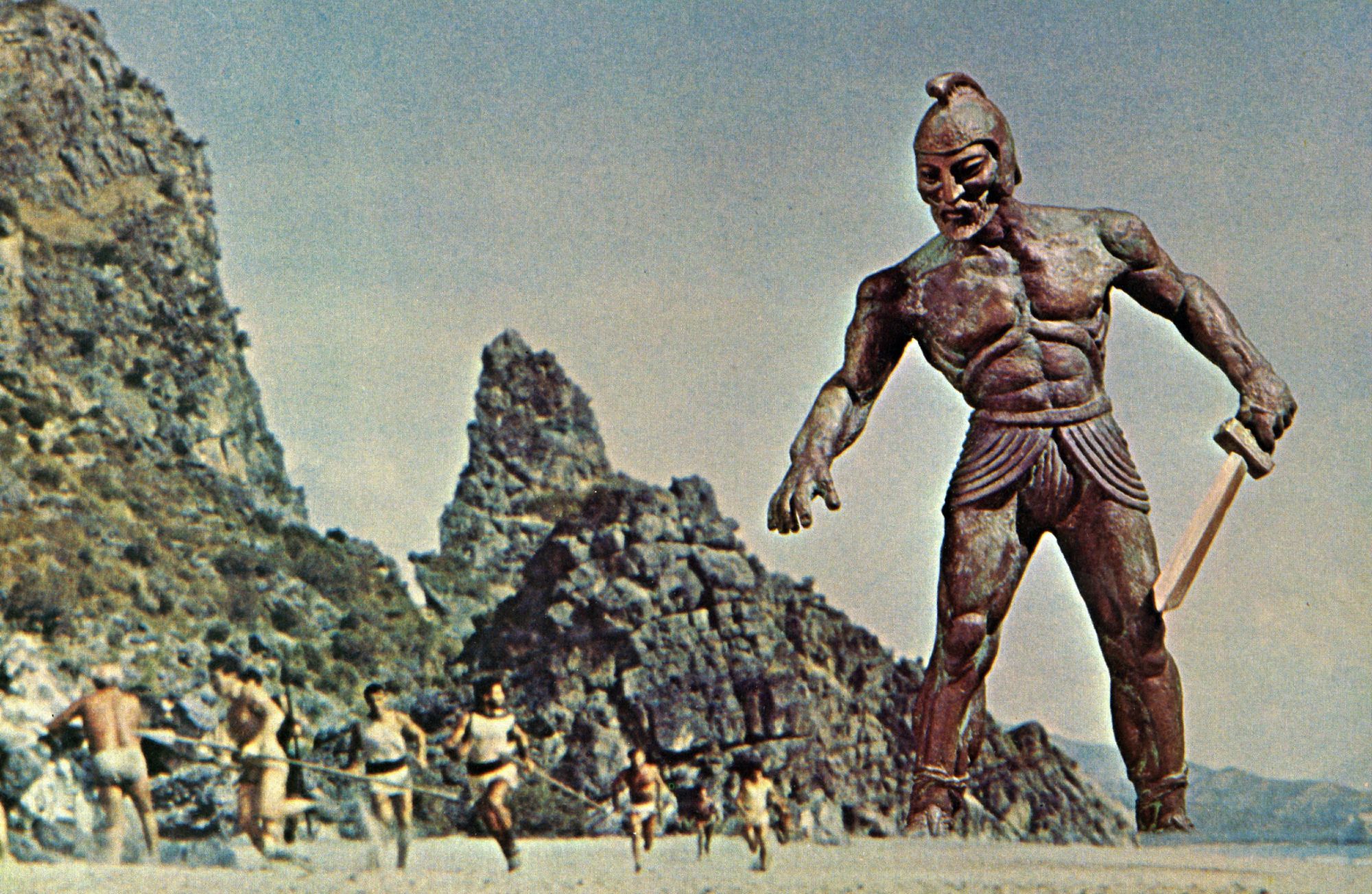 A giant, animated bronze statue of a warrior looms in the distance on a sandy beach. Sword out, the statue menaces a group of Argonauts running toward us.