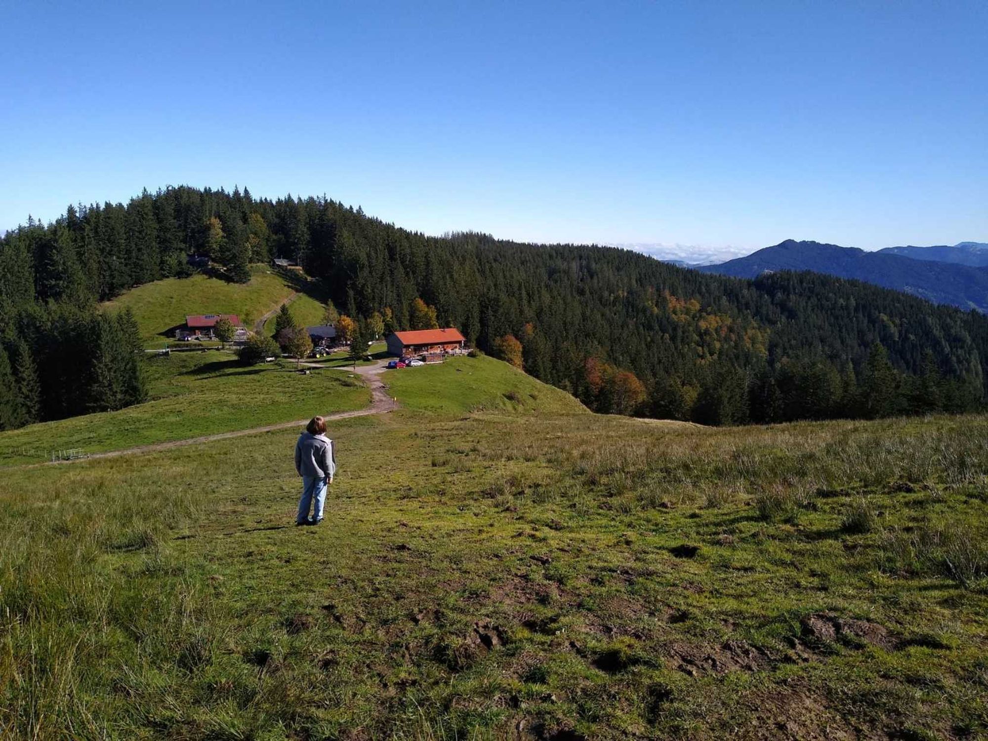 Blick über die drei Gindlalmen Richtung Schliersee. Vorne links meine Tochter schräg von hinten auf einer Wiese, rechts bewaldete Voralpen, der Himmel ist wolkenlos blau.