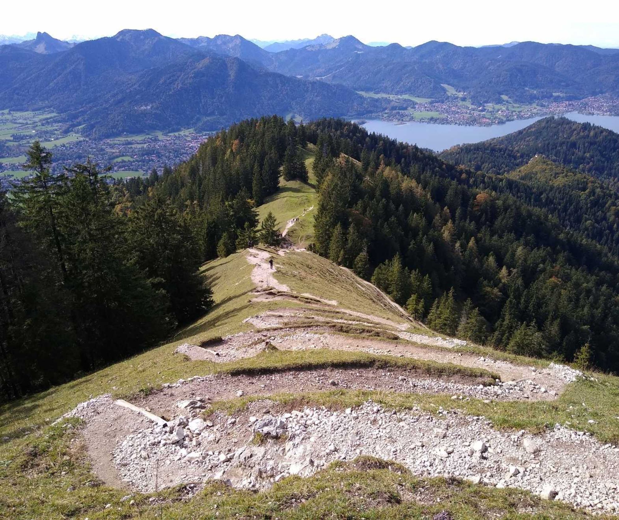 Blick vom der Baumgartenschneid hinab Richtung Tegernsee: der Wanderweg läuft in stellen Serpentinen hinab, rechts und links Bäume in der Ferne der Tegernsee und angrenzende Gebirge.