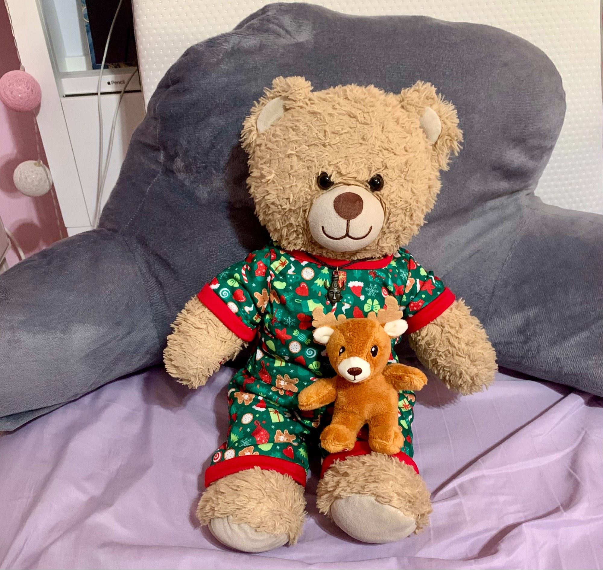 A teddy bear in Christmas patterned pajamas holds a tiny plush reindeer on his lap.