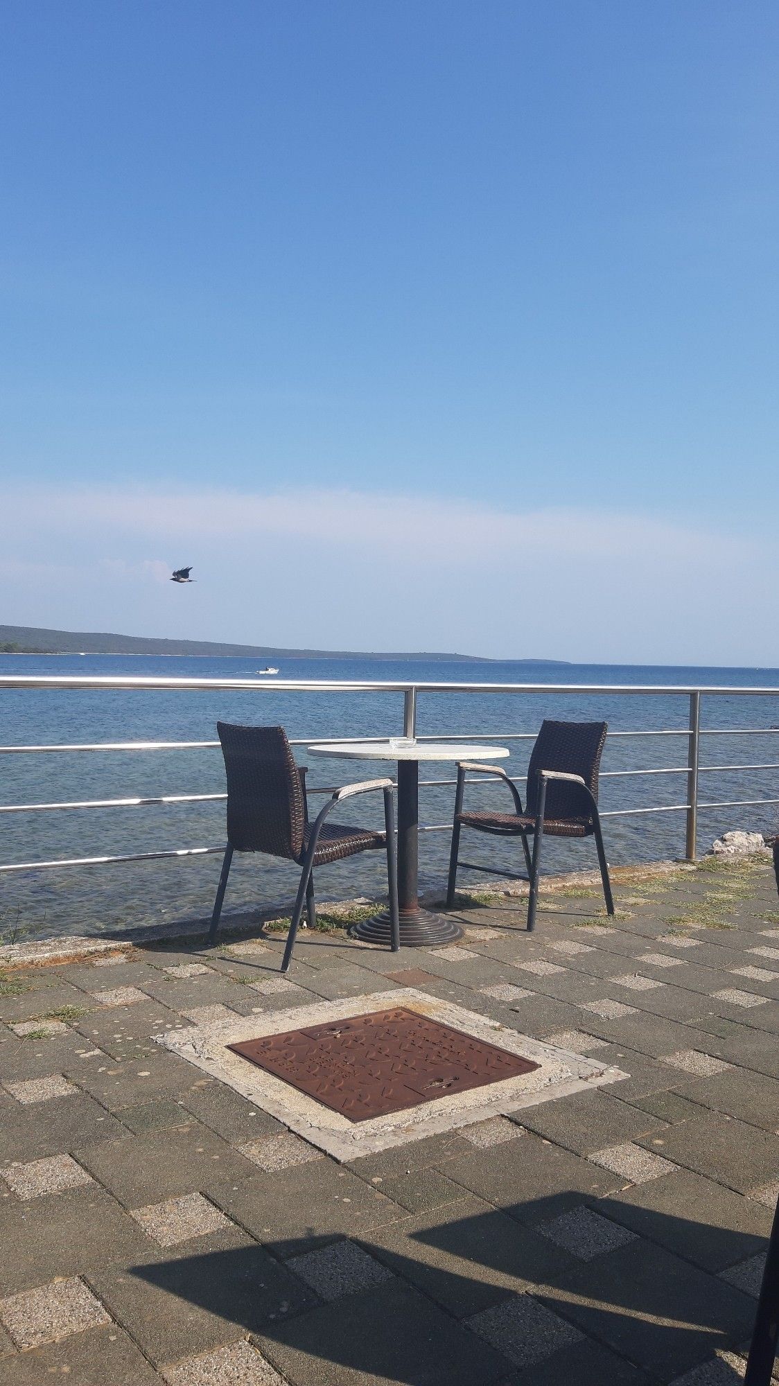 Zwei heflechtbezogene Stühle an Metalltisch, auf Waschbetonplatten vor glänzendem Nirozaun vor Meer vor Insel unter blsuem Himmel. Krähe fliegt vorbei, zwei eher unscharfe Boote.