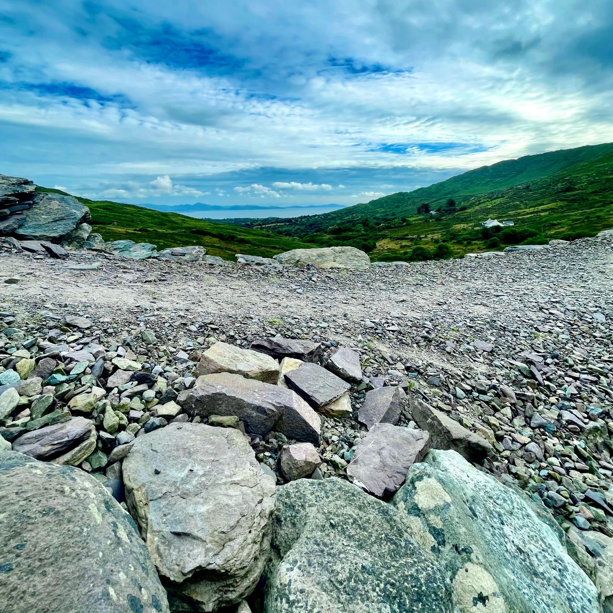 The rocky, gravel top of the thick wall of the ring fort looking out on a hilly landscape under a partly cloudy sky with green vegetation. In the distance a clear view of the Atlantic sea and any incoming seaborne threat.