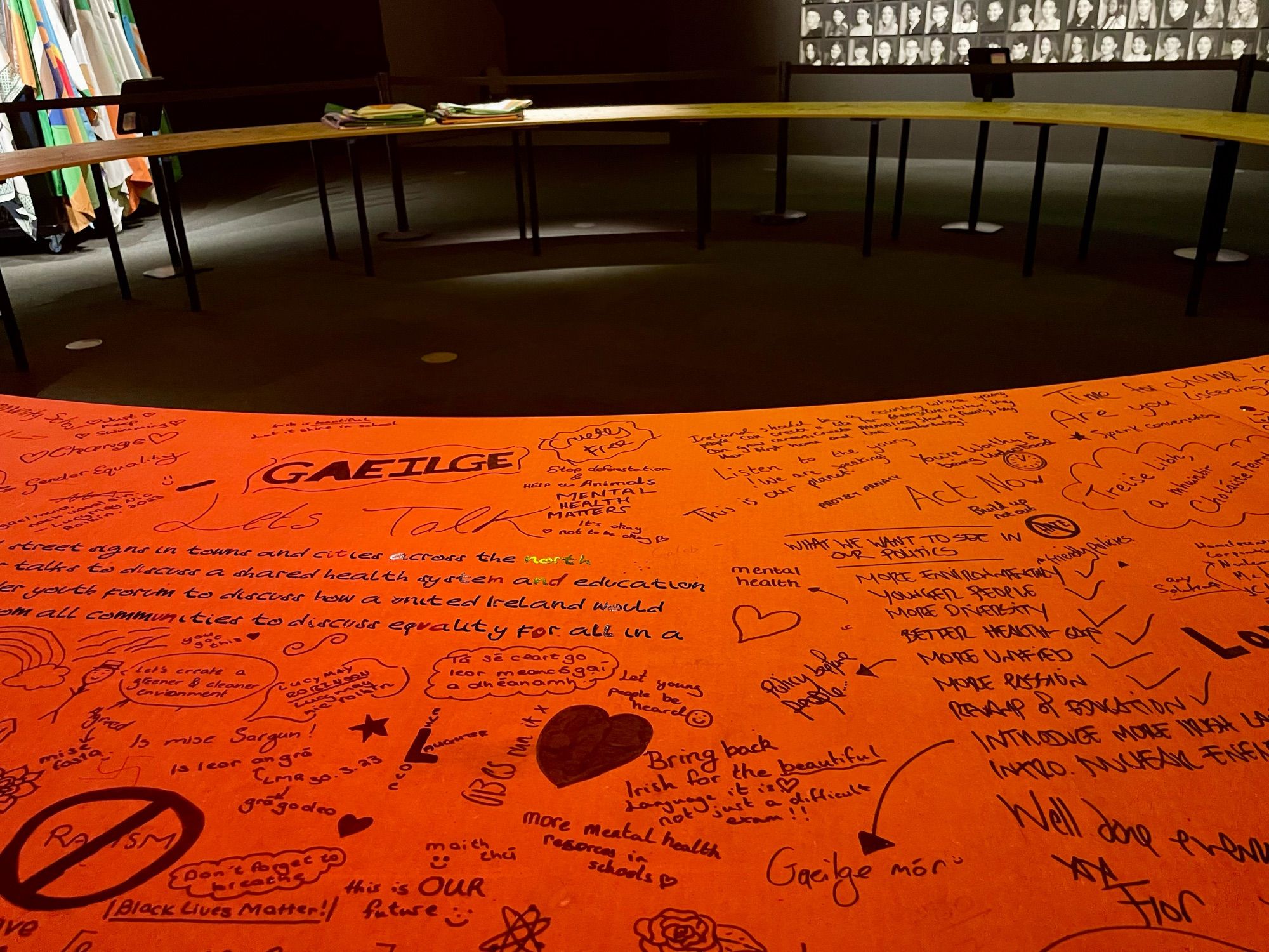 A large round table orange-lit with various handwritten graffiti messages, ideas, and doodles in different styles made by black Sharpie. The background shows flags, and a wall with numerous small portraits.