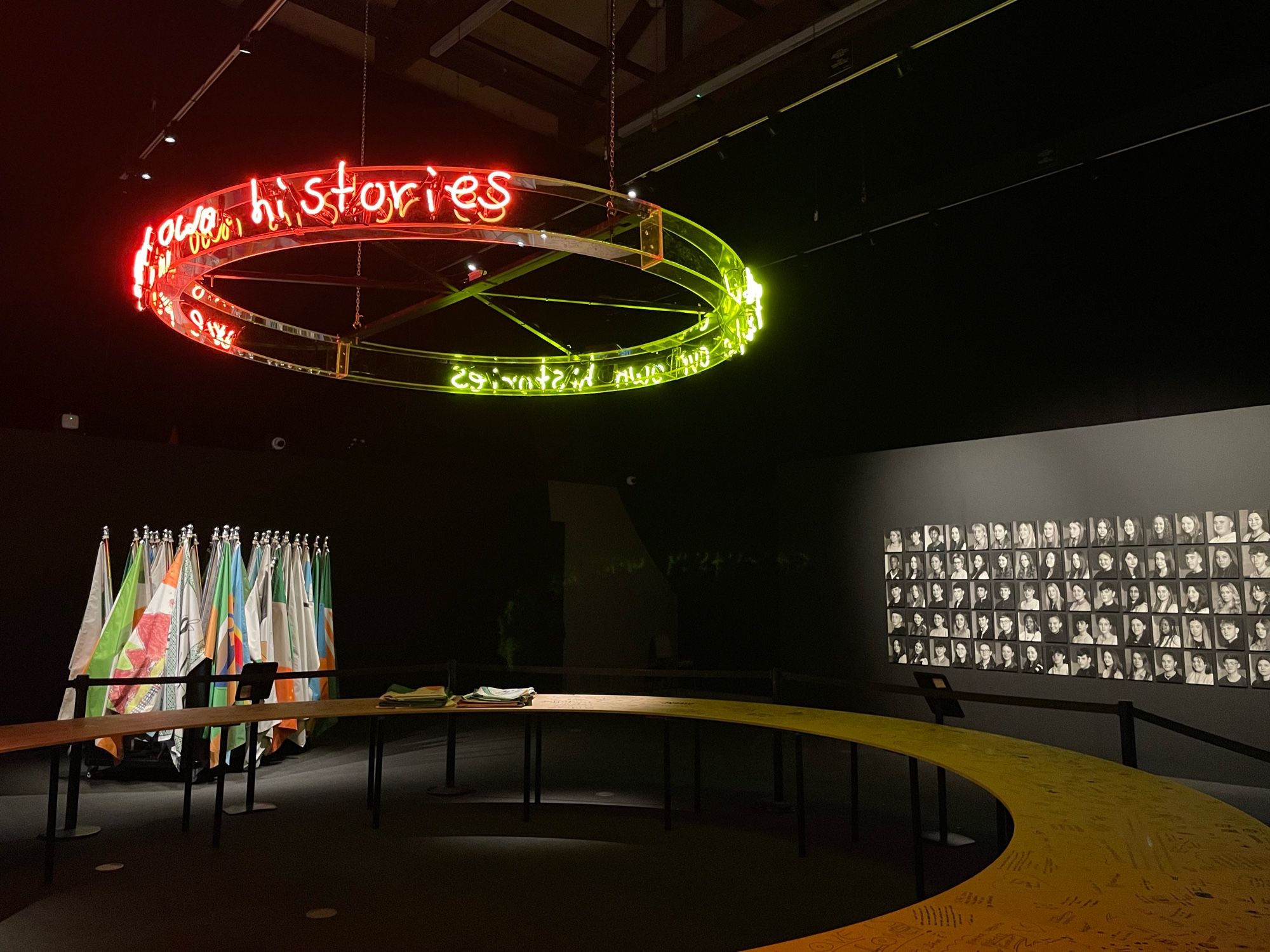 An art exhibit featuring a circular neon sign with the text "your histories" in red and green, a display of colorful flags on poles, and a wall with a grid of black and white portraits.