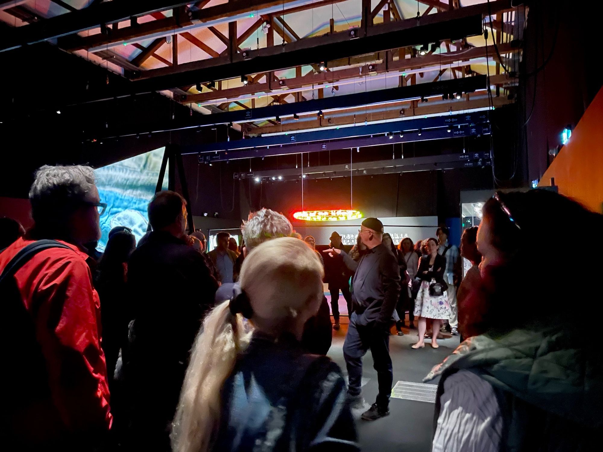 A group of people gathered inside a dimly lit room with a speaker addressing them. A large screen and neon sign are visible in the background. The ceiling displays an exposed beam structure.