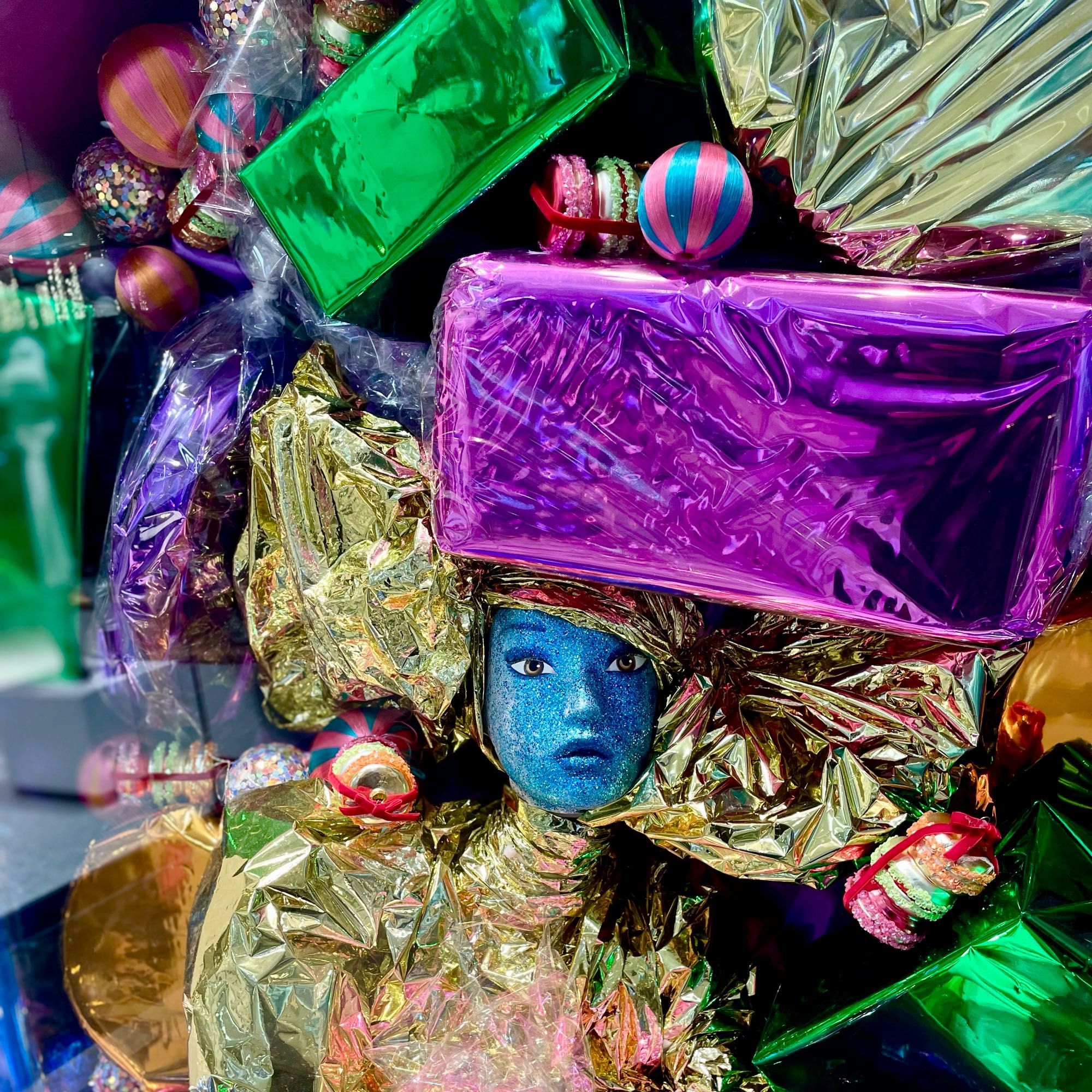 Blue mannequin in gold foil under avalanche of colourful wrapped presents.