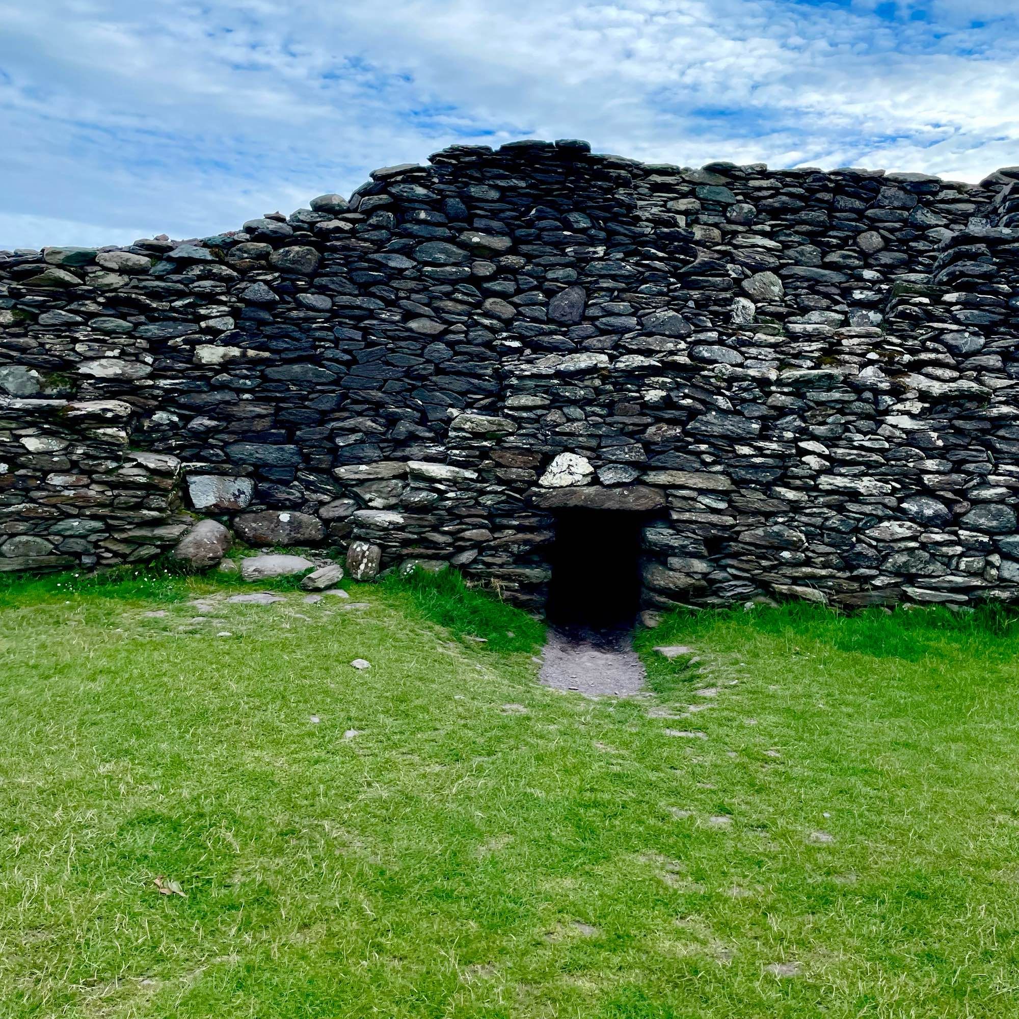 Ancient stone wall with a small, dark entrance at the base and green grass in the foreground. Blue sky with clouds in the background