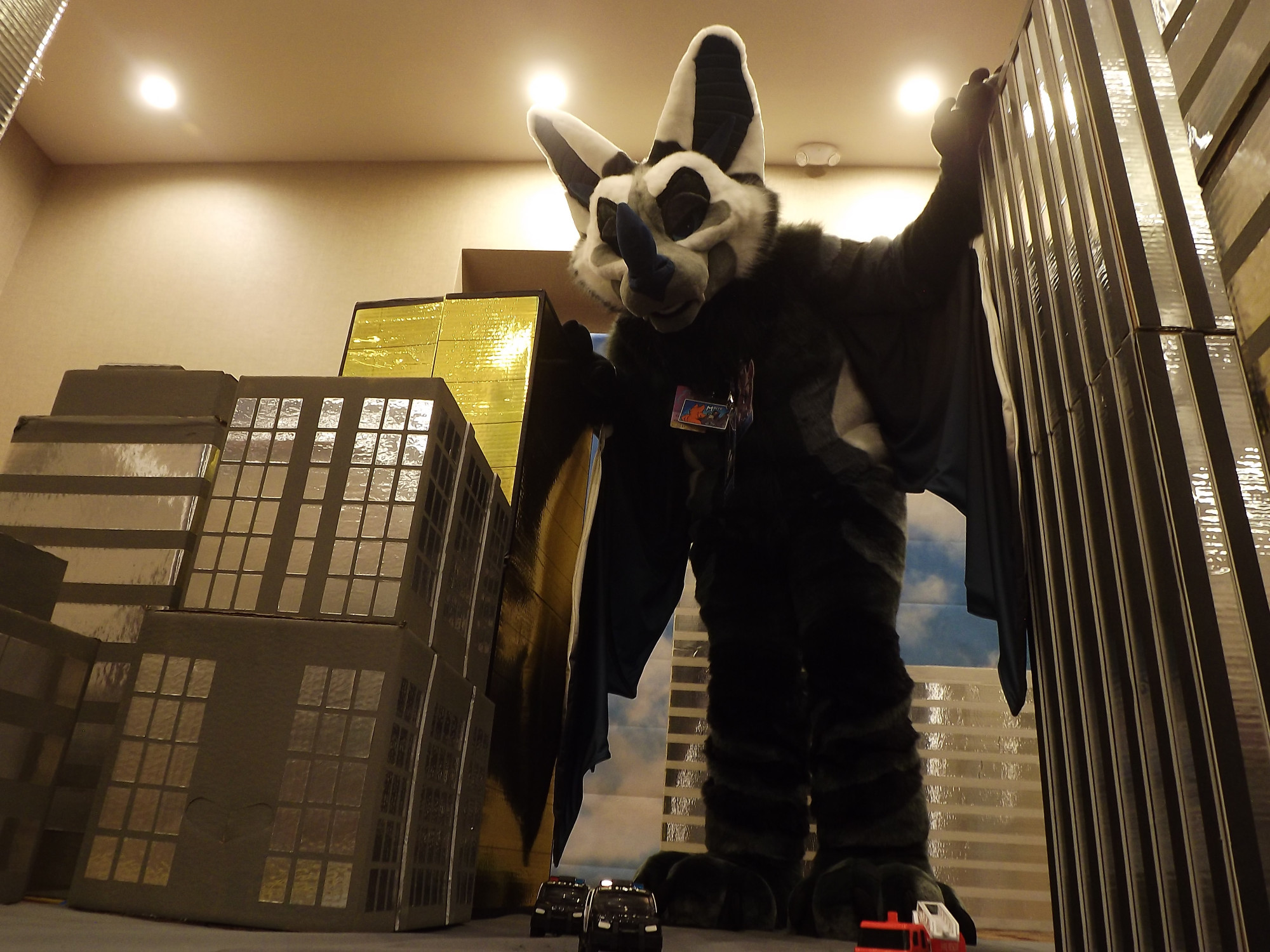 A bat fursuit leaning on some fake buildings in a prop city at a macro meetup implying they are gigantic in the city. 