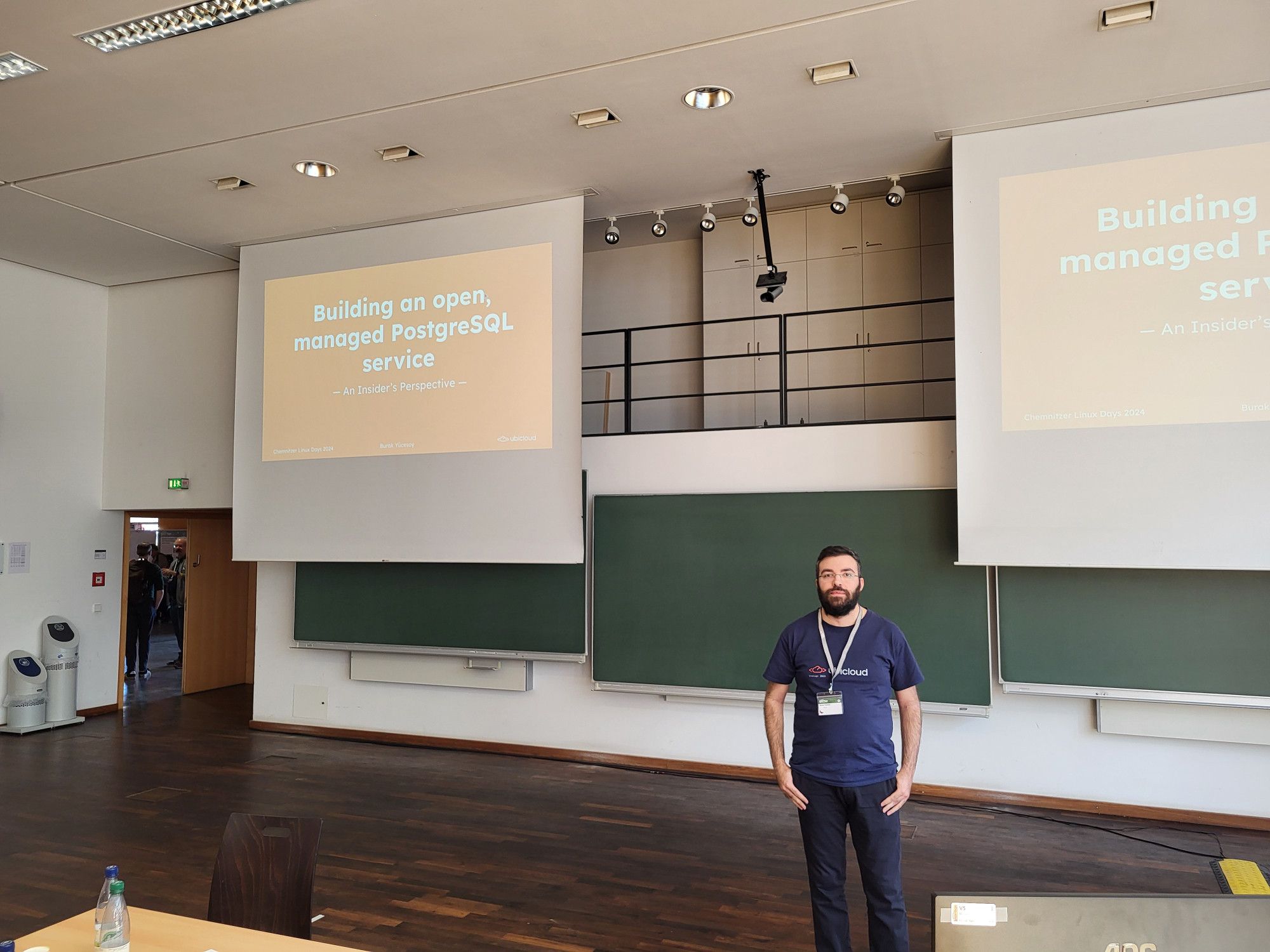 Burak Yucesoy standing in front of the audience, in the background the slides for his "Building an open managed PostgreSQL service" talk are seen on the screen