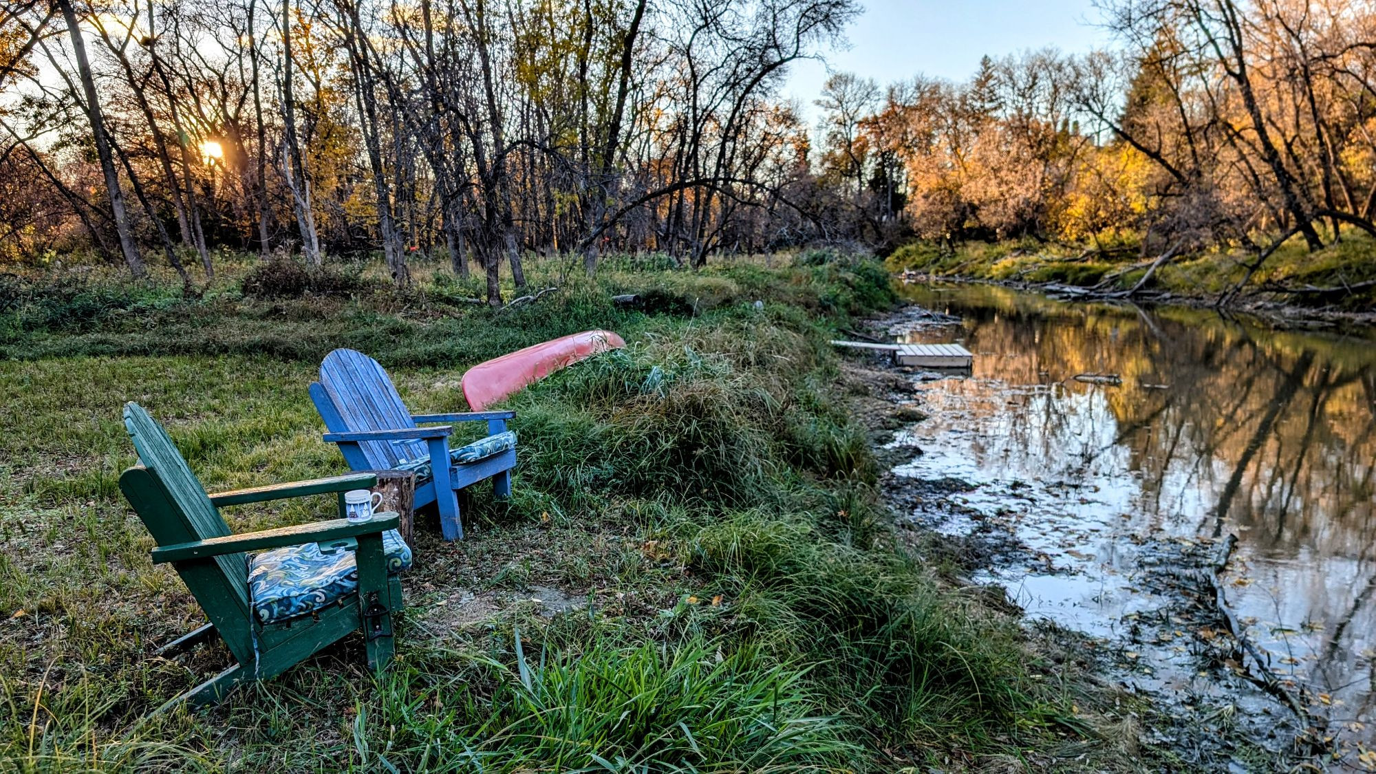 The setting sun shining over a clearing beside a little Forest River with two wooden chairs in one painted blue and one painted green. A red canoe sits waiting on the bank beside a little dock in the river. 