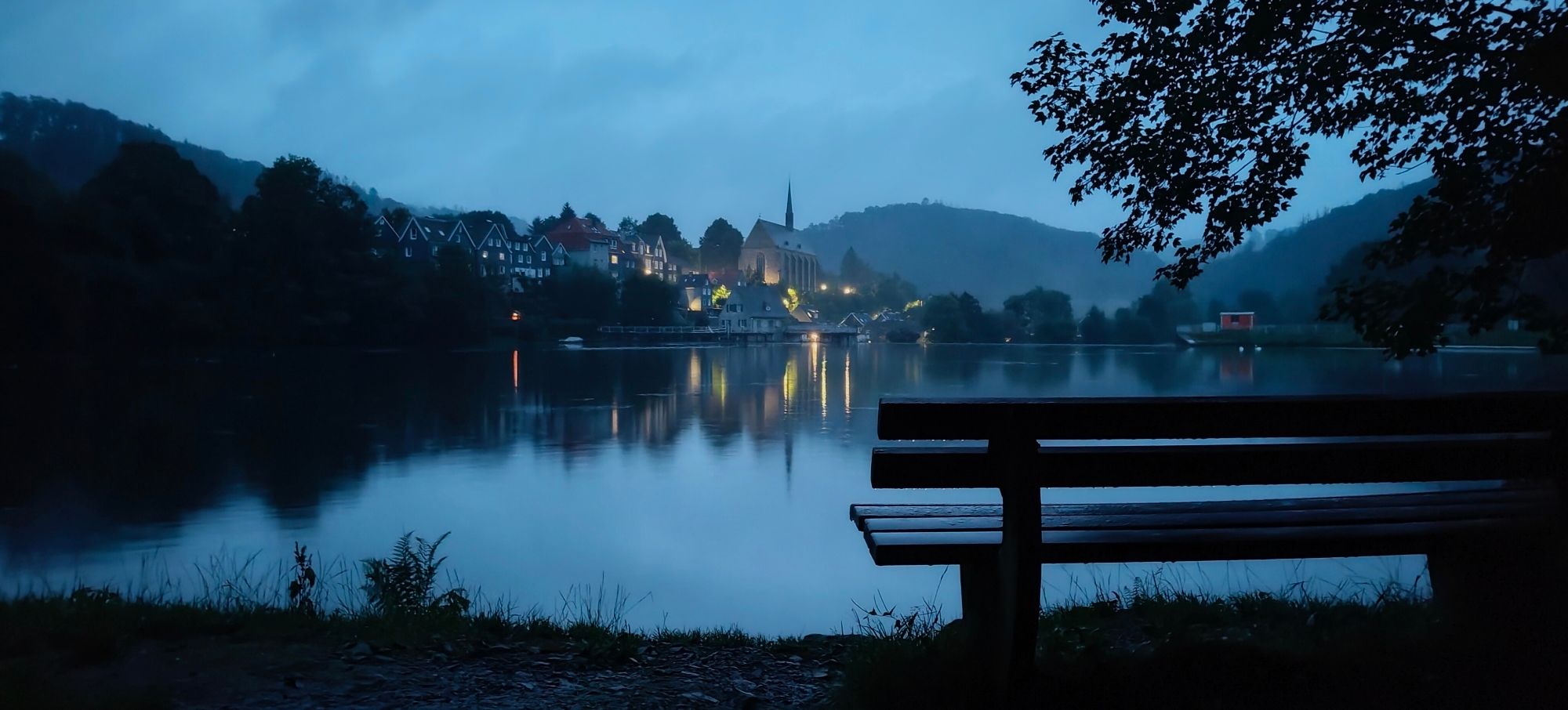 Ein See mit einem alten Dorf am Horizont und einer Parkbank im Vordergrund. Es ist später Abend.