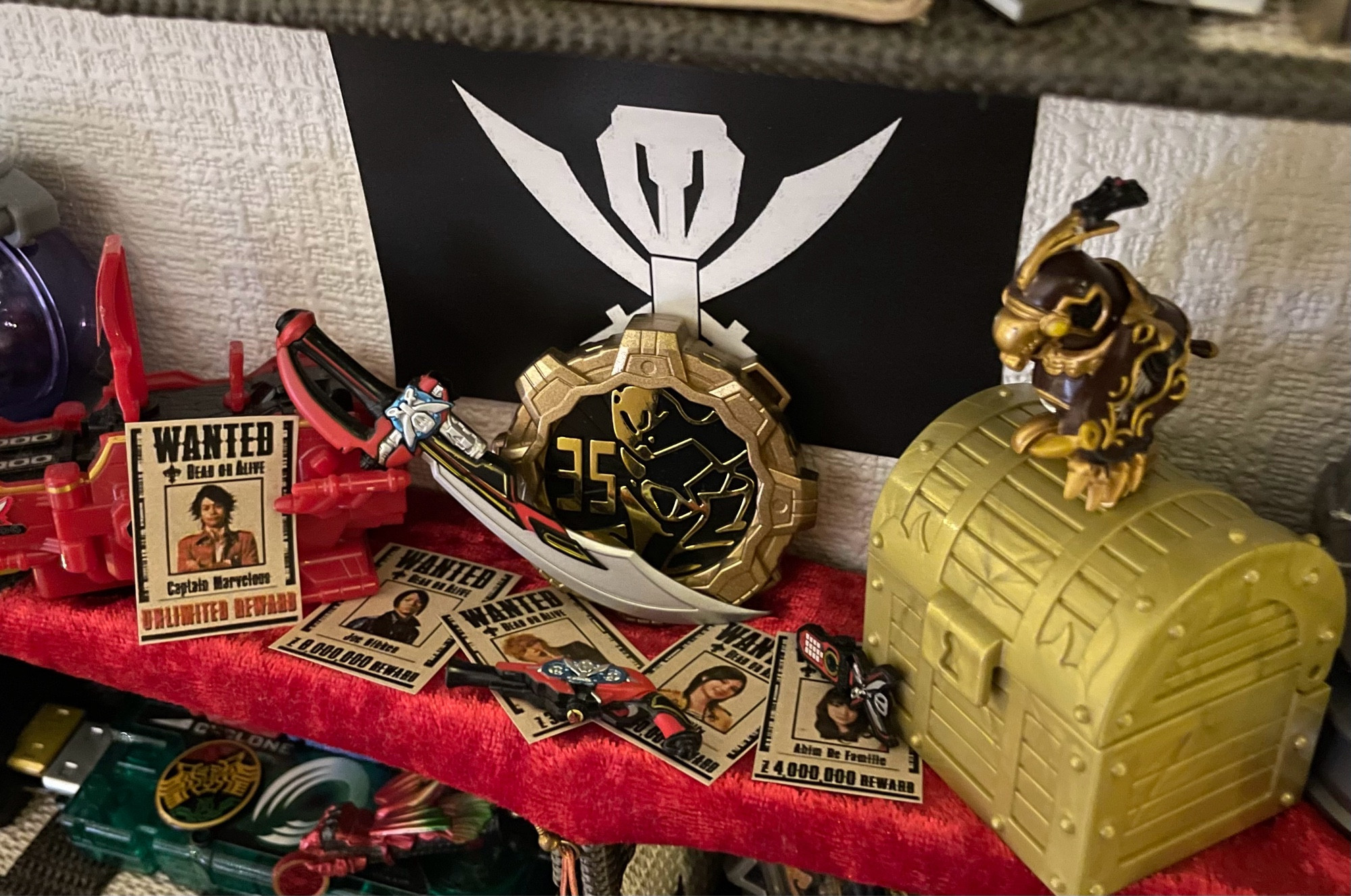 a miniature gokaiger display on red velvet featuring the gokai galleon, a gold treasure chest with navi perched on top, a gokaiger flag pinned to the wall, the 35 gear from zenkaiger, and tiny wanted posters with a tiny gokai saber, gun, and phone scattered on top