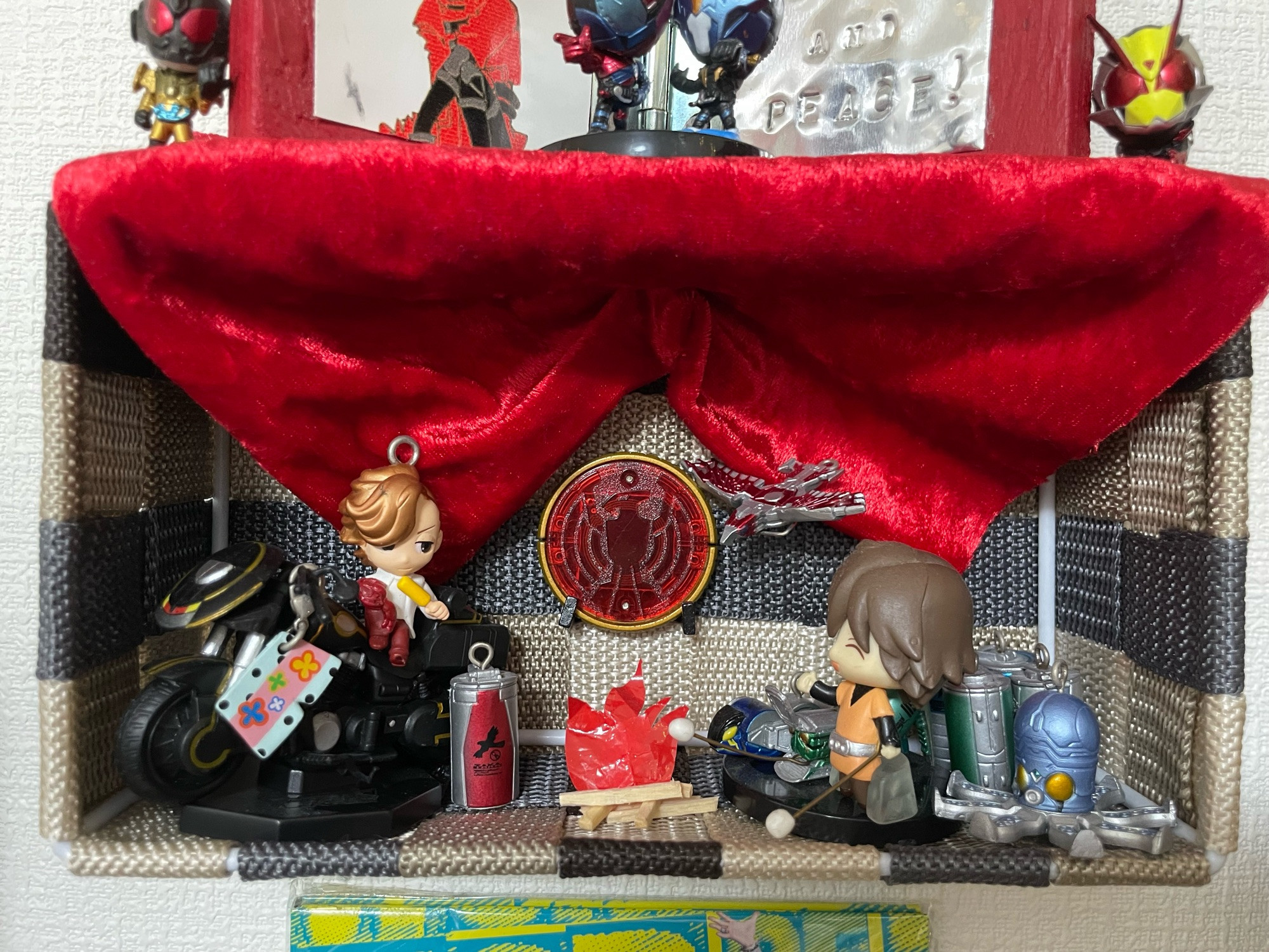 a miniature kamen rider ooo display that I totally didn't make to spite the 10th anniversary movie, featuring ankh on the bike with boxers dangling from the handlebars, a little fire made of sticks and plastic, and eiji and some candroids toasting marshallows. above them is a canopy of red velvet with the tajador medal pinned to back underneath it