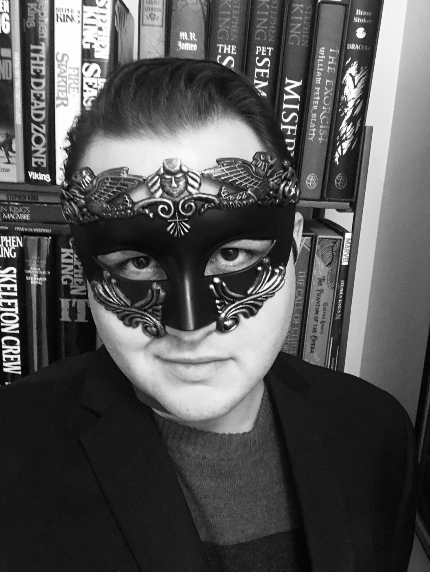 Alt text: me staring at camera with a slight smile, and I’m wearing a black gothic mask with gold edging, though the photo is in black and white so it looks silver