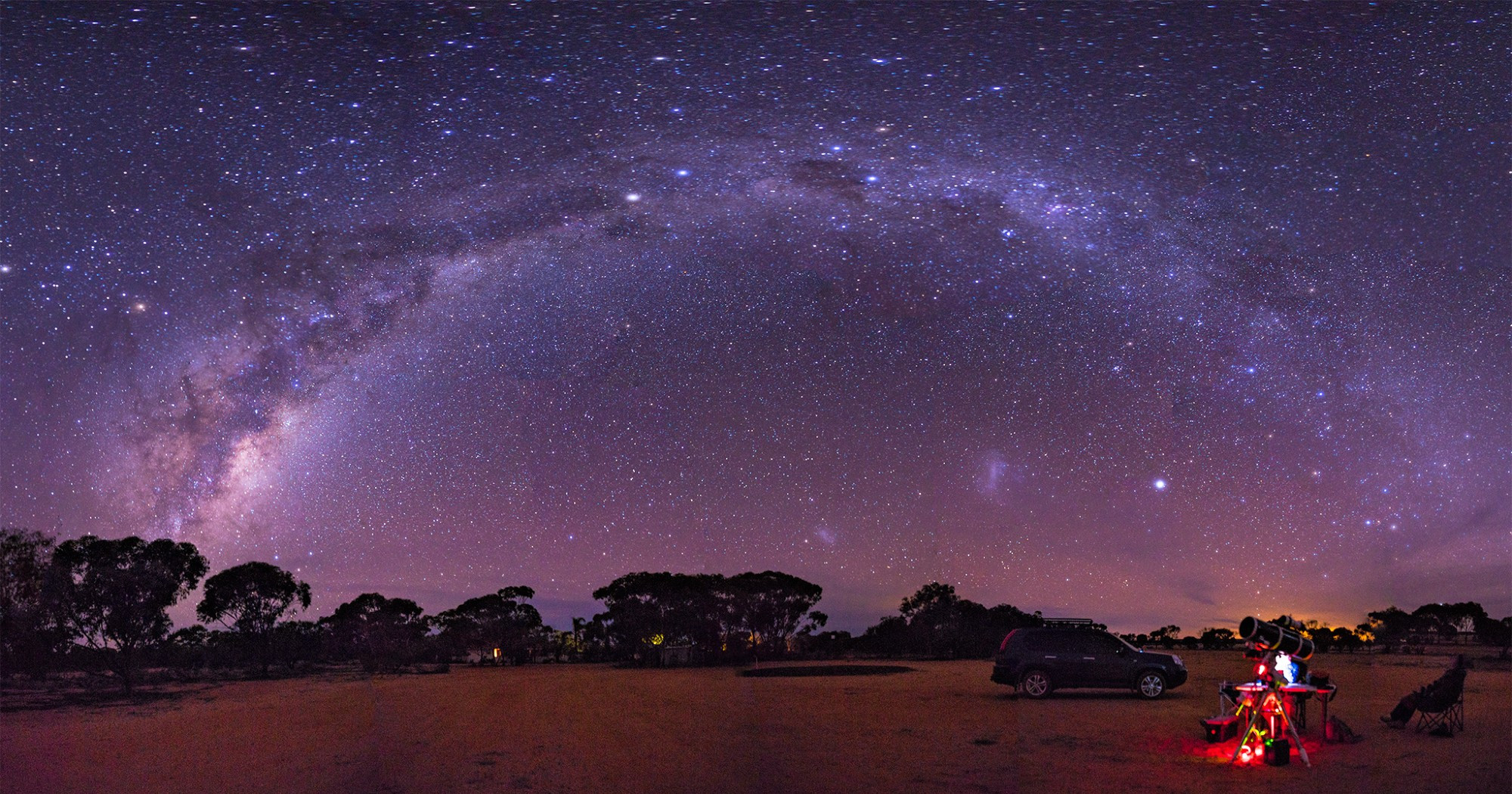 a merged and stitched panoramic view of the Milky way in an arch over a dirt golf course in the West Australian wheatbelt with a deep sky telescope setup illuminated in the foreground