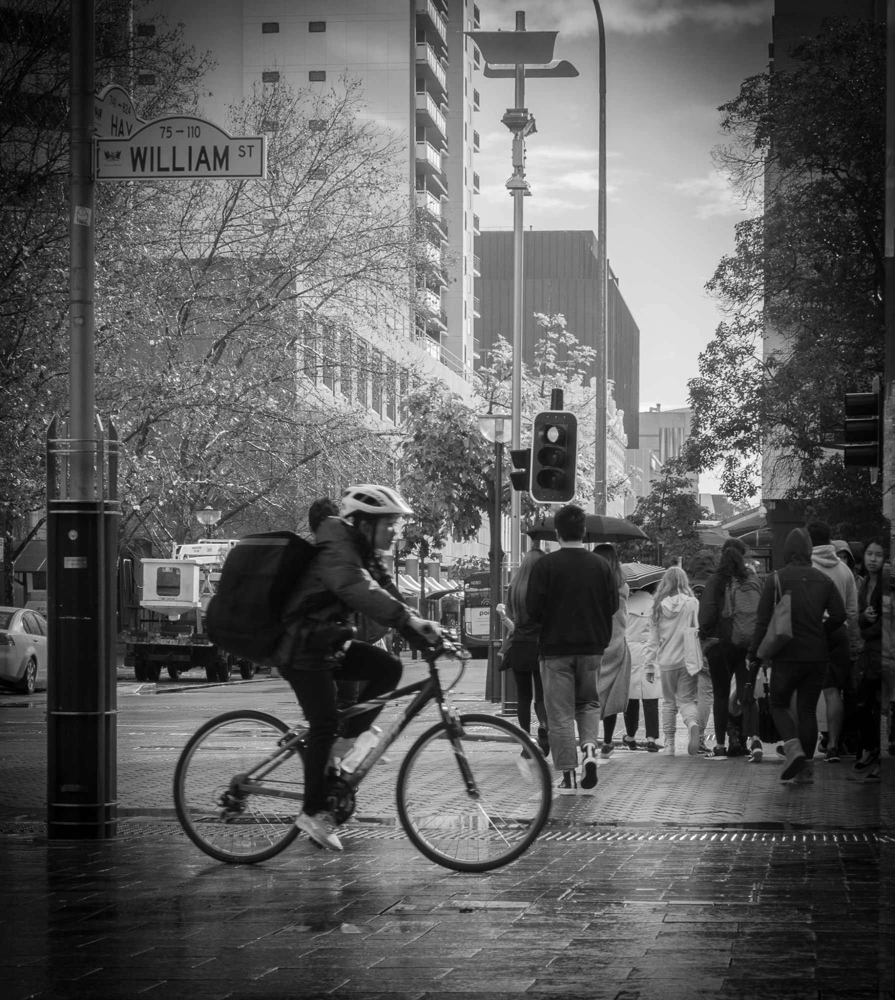 A black and white street photography style photo of a delivery rider cyclist passing behind a group of people crossing the street