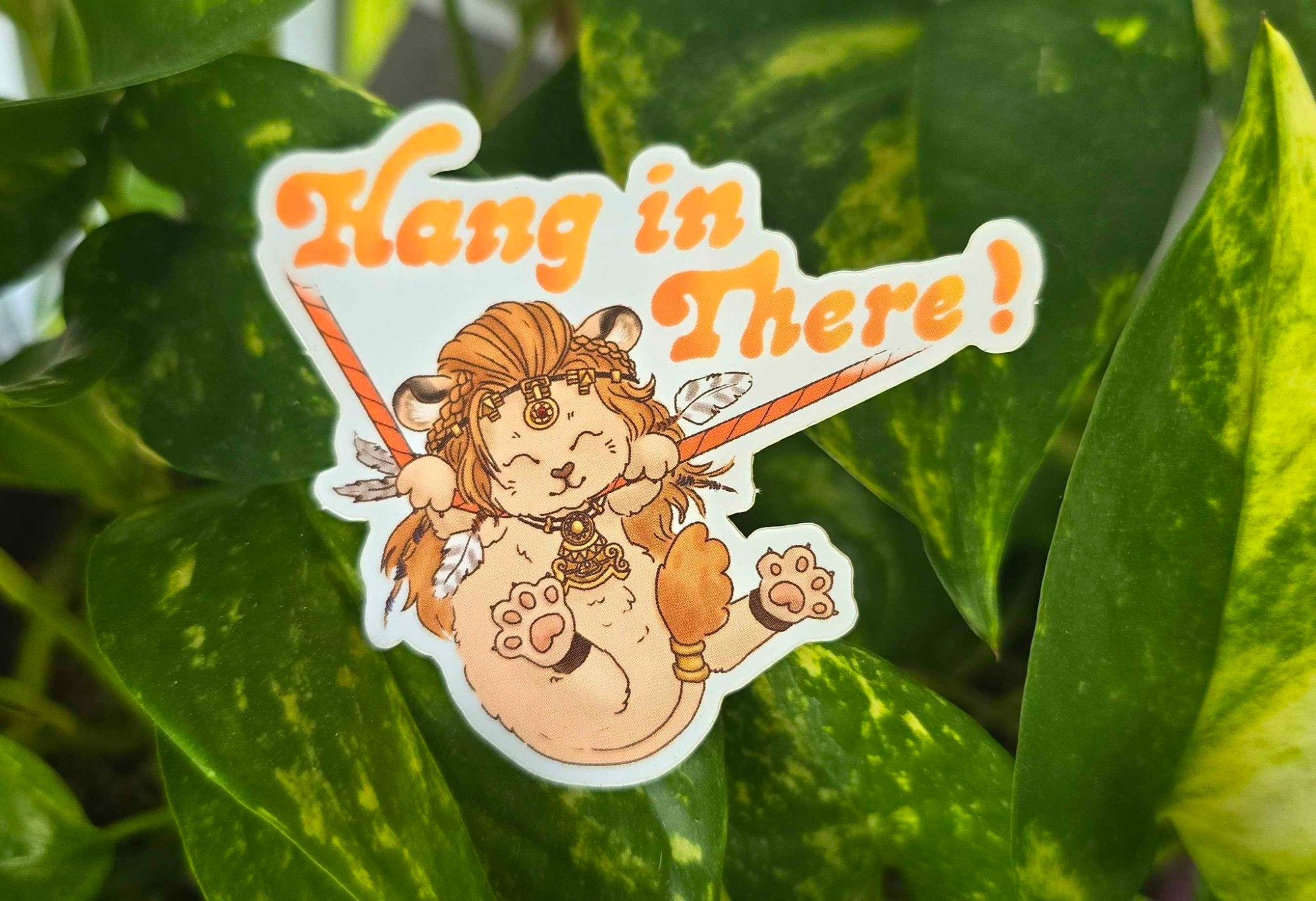 Sticker of Wuk Lamat as a cute little kitty, hanging from a line like the inspirational cat poster, with the words "Hang in There!" above her in an orange and yellow gradiated 70s font.