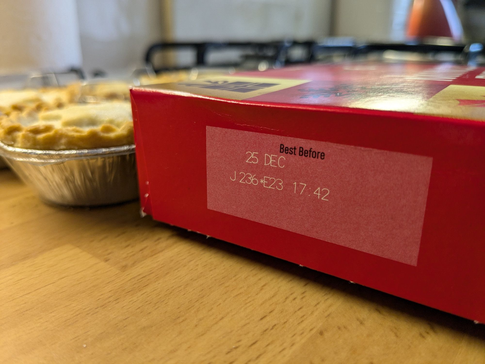 On a kitchen surface, the close-up of the end panel of a predominantly red-packaging box of mince pies, showing a "Best Before" date of "25 DEC" with some coding. Next to the box are the tray of mince pies from within it.