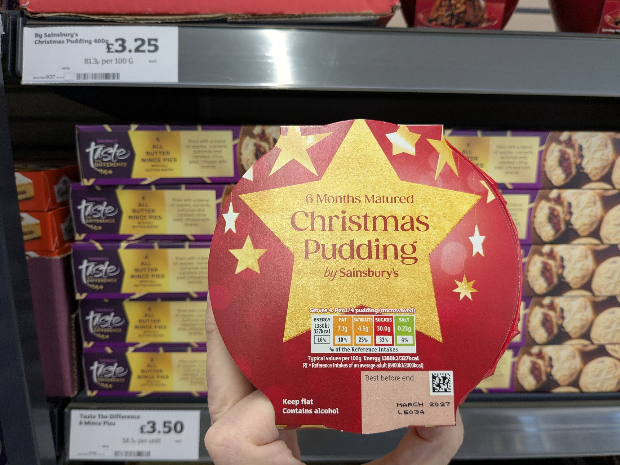A packaged Christmas pudding, held in front of a shelf of mince pies.