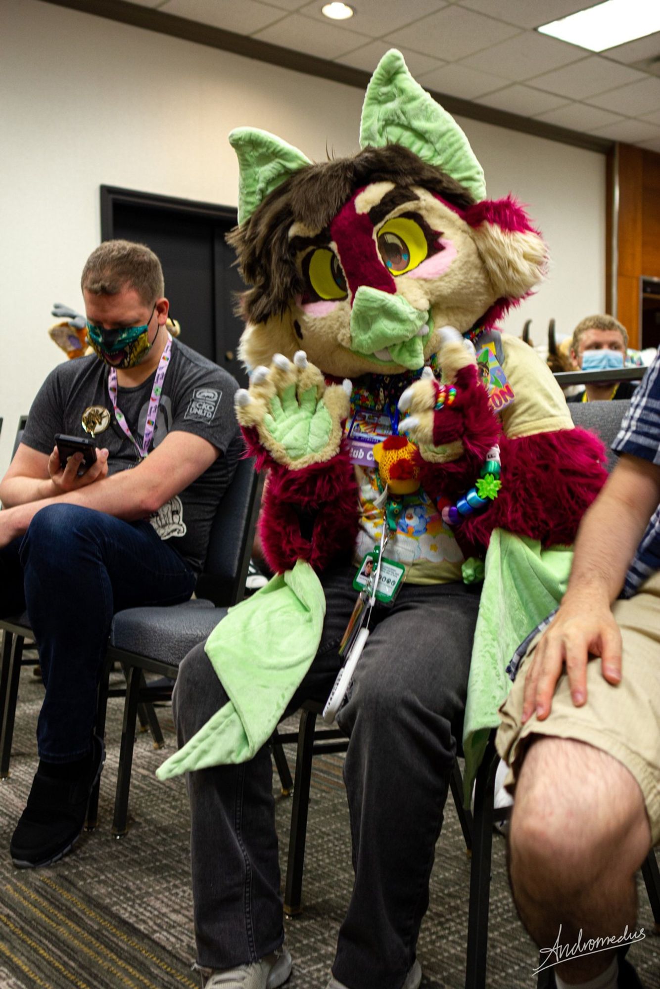 A bat fursuiter with red, pink and tan fur, brown hair and eyes, yellow sclera, and green skin sitting on a chair in a conference room and waving at the camera. He is wearing a collar with various badges hanging from it, a yellow Care Bears shirt, a lanyard with a fan attached to it, and a pair of dark grey jeans