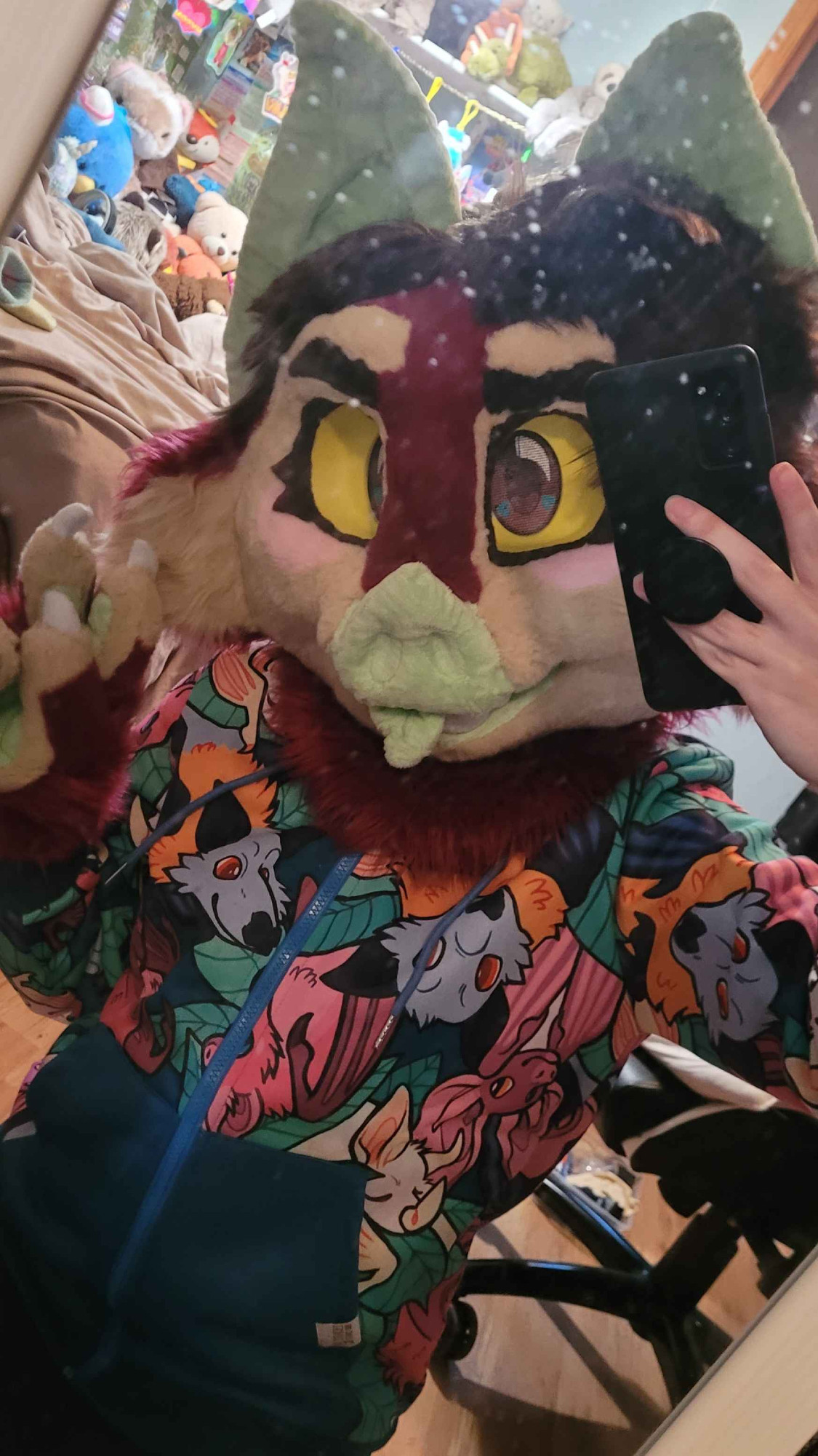 A bat fursuiter with short, brown hair and furrowed eyebrows taking a mirror selfie. He is wearing a sweater covered in bats in leaves