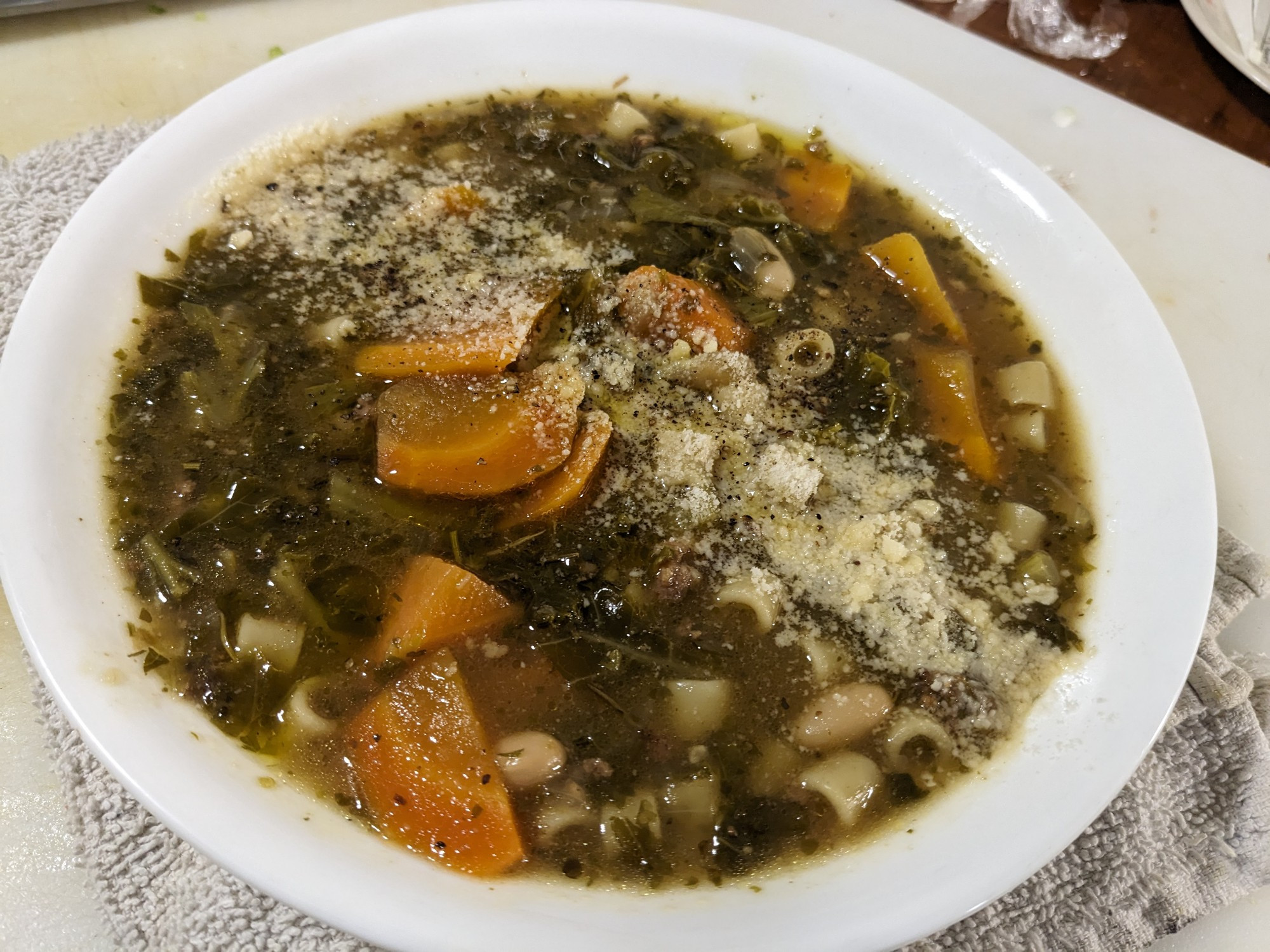 A wide, low, white ceramic bowl, filled with a healthy portion of sausage and kale soup, ditalini pasta, white beans, and carrot obliques in the rich herbed broth. A stripe of grated Parmesan garnishes the middle of the bowl, complimented by a drizzle of green extra virgin olive oil and fresh cracked black pepper.