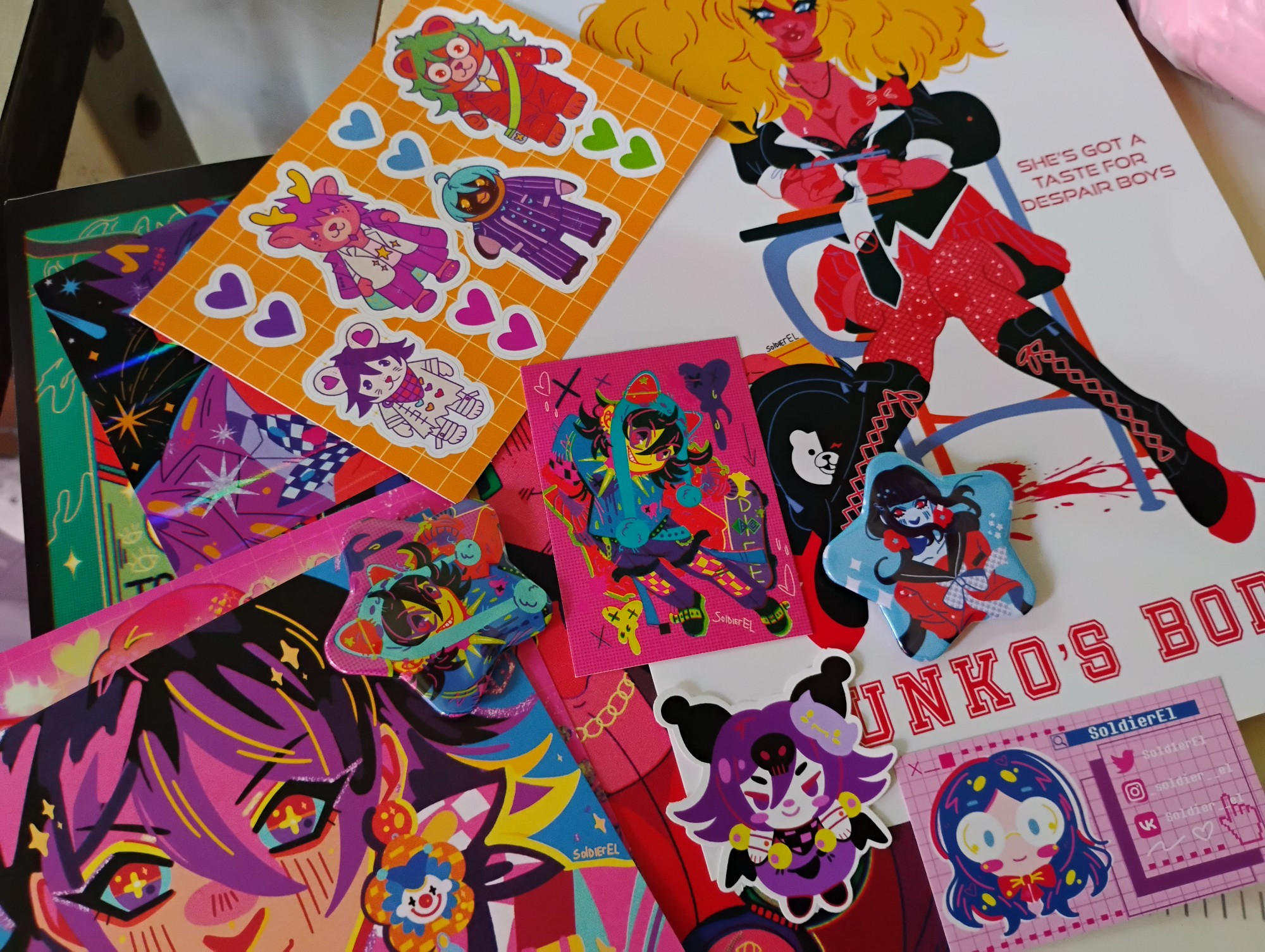 Photo featuring diverse merch of artist soldier el, including a jennifers body themes junko enoshima print, two fashionable kokichi prints, kuromi kokichi sticker, half covered kaito momota print, kokichi ouma star button and maki star button, as well as sylvanian danganronpa v3 sticker sheet with bear gonta, deer kaito, penguin shuichi and mouse kokichi