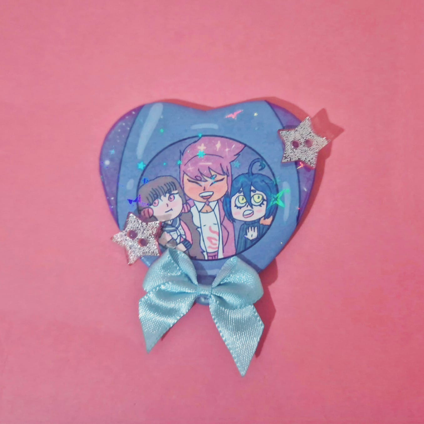 botton de coração com um laço azul e dois botões de estrela de glitter rosa pink. O bottom contém um foguete e na janela dele podem ser vistos os personagens kaito momota, maki harukawa e shuichi saihara