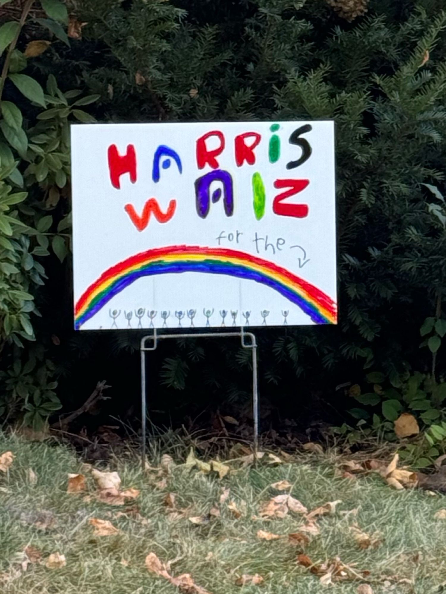 A handmade political sign with a rainbow that says “Harris Walz” and then in smaller letters below “for the” and an arrow pointing to a rainbow with several stick  people standing under it