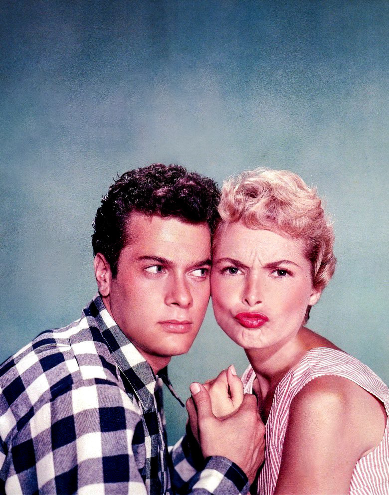 Tony in a checkered blue and white collared shirt, Janet in a red and white striped dress, they are sitting cheek to cheek with their hands clasped together. Janet is making a weird kissy face toward the camera. 